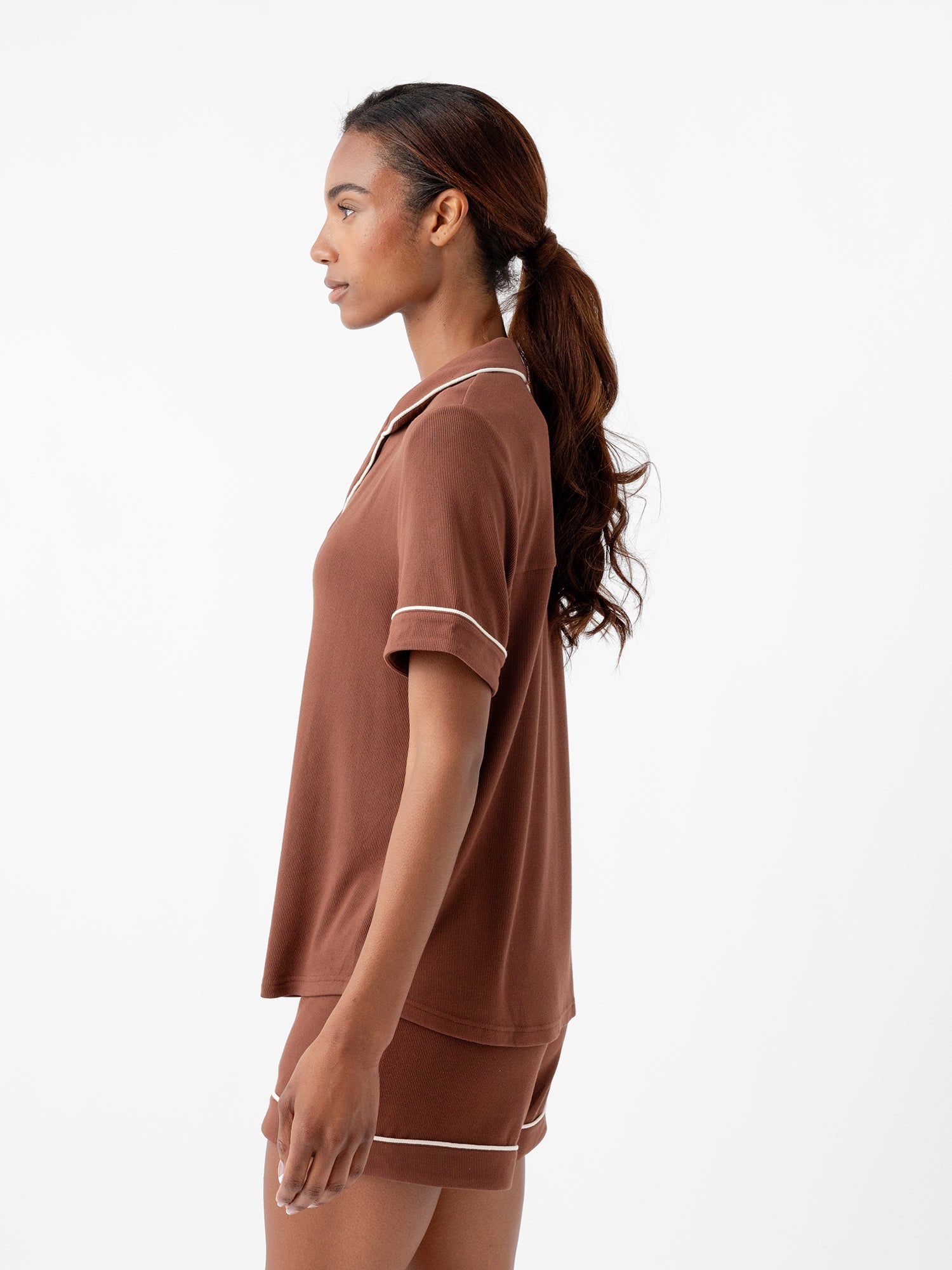 A person with long, wavy hair tied back in a ponytail is standing in profile, wearing Cozy Earth's Women's Bamboo Rib Knit Classic Short Sleeve Pajama Top and matching shorts with white trim. The background is plain white. 