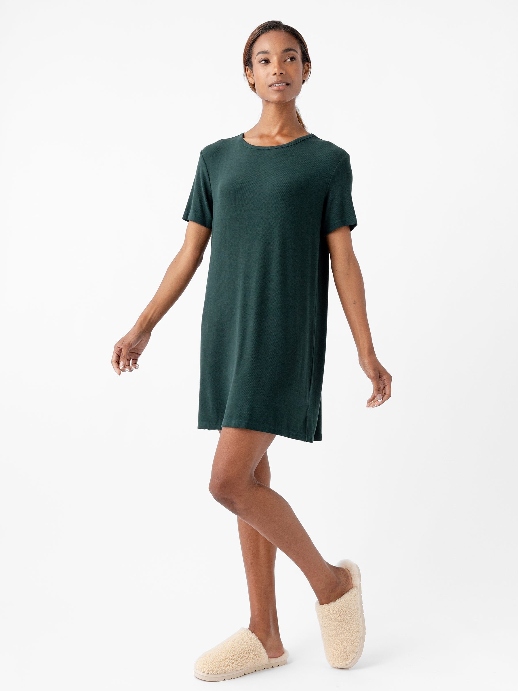 A person is standing against a plain white background, wearing a dark green *Women’s Bamboo Rib-Knit Boyfriend Sleep Dress* by *Cozy Earth*, paired with light beige, fluffy slippers. The person has one leg slightly crossed over the other and has a relaxed expression on their face. 