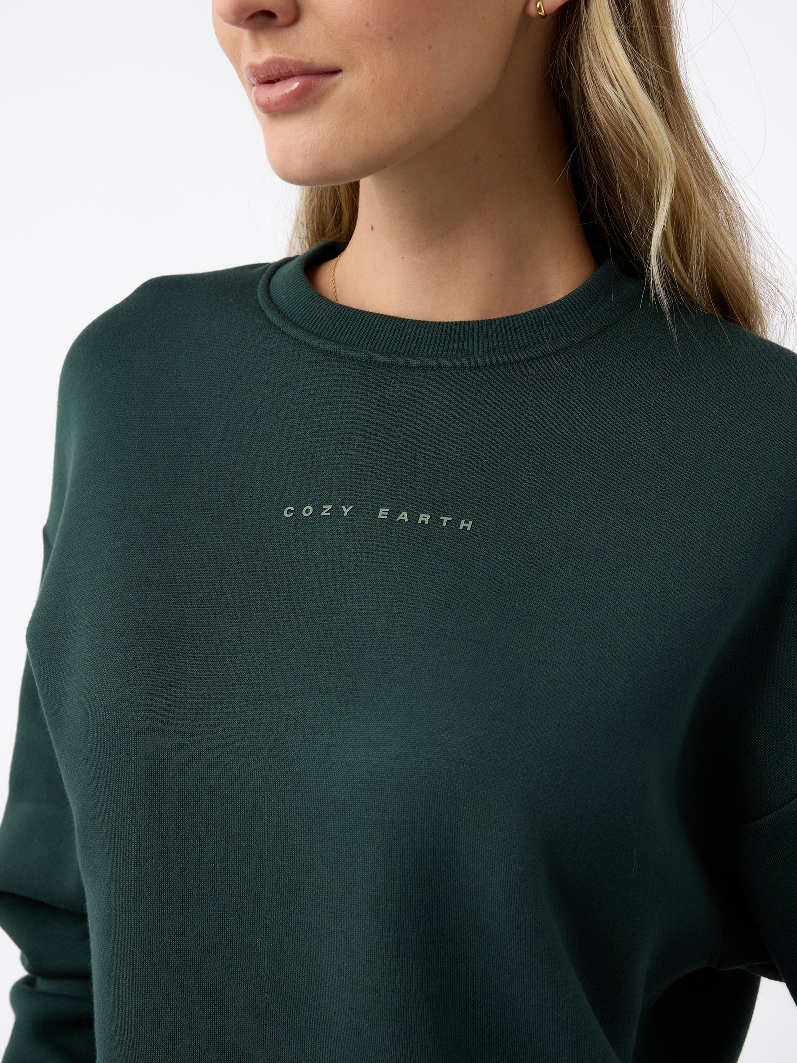 A person wearing a Women's CityScape Crewneck sweatshirt in dark green from Cozy Earth. The individual, with long, light-colored hair, is facing slightly to the side against a plain background. 