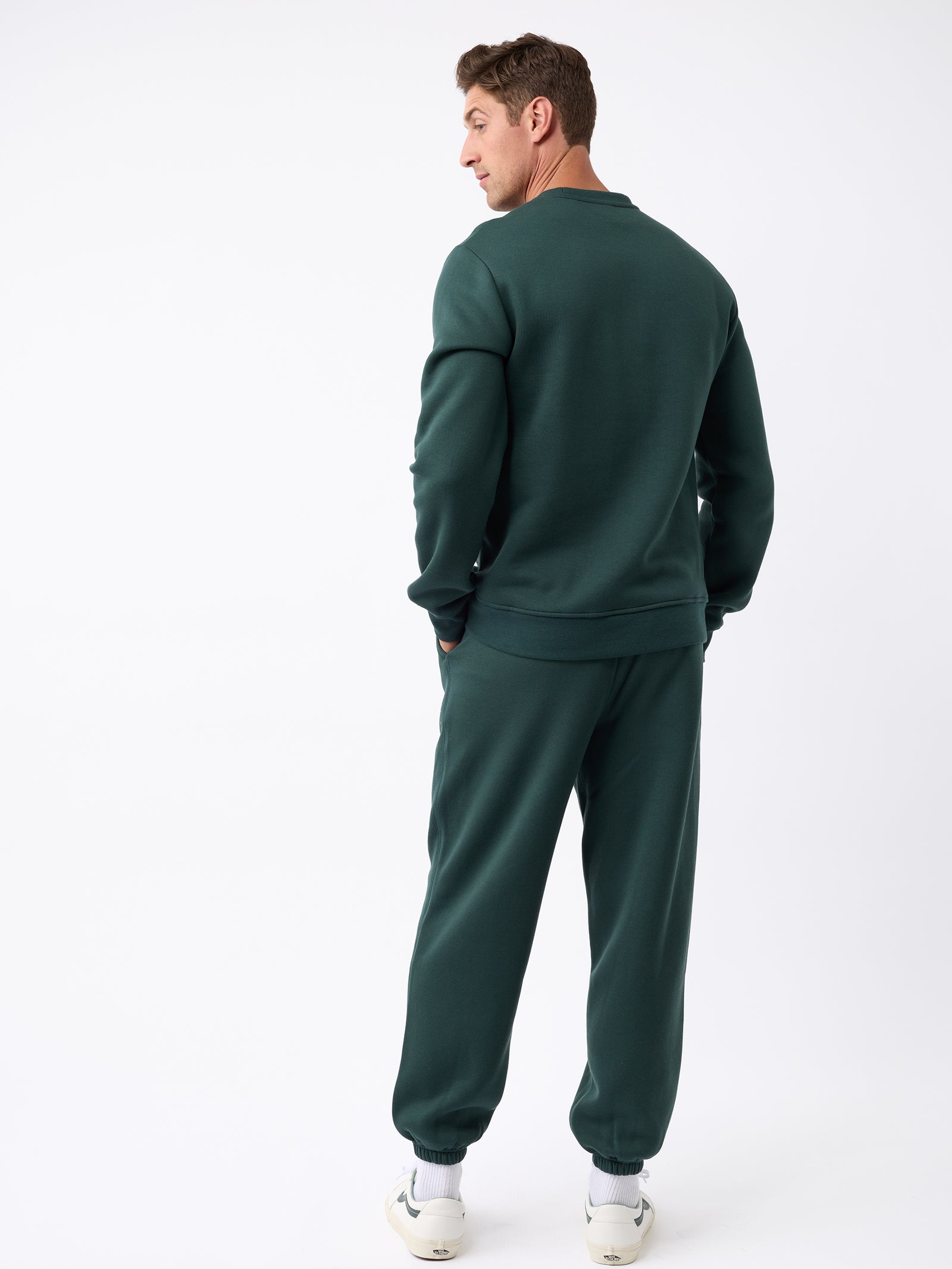 A man, with short brown hair, stands facing slightly away from the camera in a white studio setting while wearing the Men's CityScape Crewneck and matching sweatpants from Cozy Earth in dark green. 