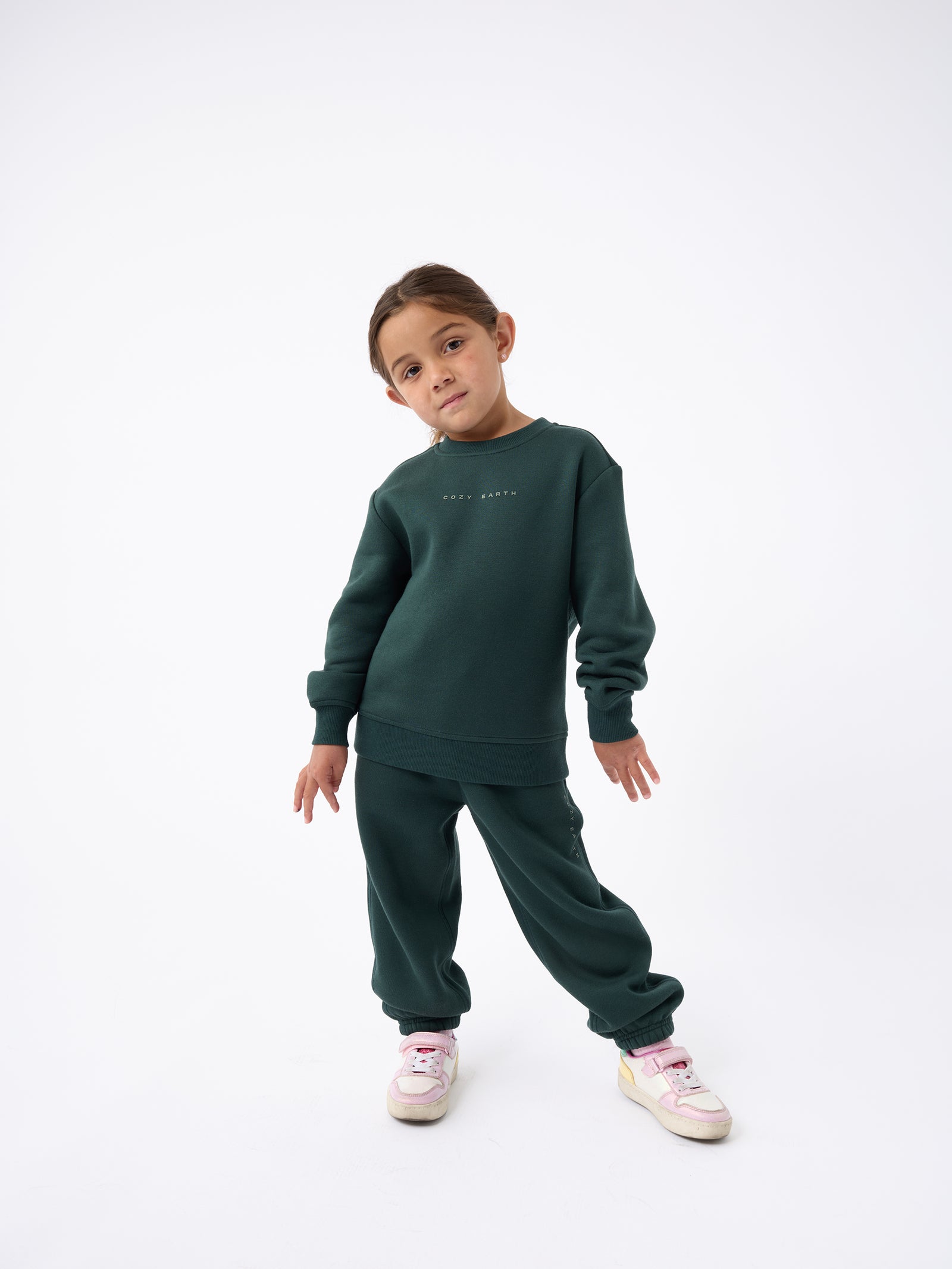 A young child poses against a white background, confidently wearing the Kid's CityScape Crewneck by Cozy Earth in dark green, paired with matching sweatpants and pink and white sneakers, standing with hands slightly bent at the sides. 