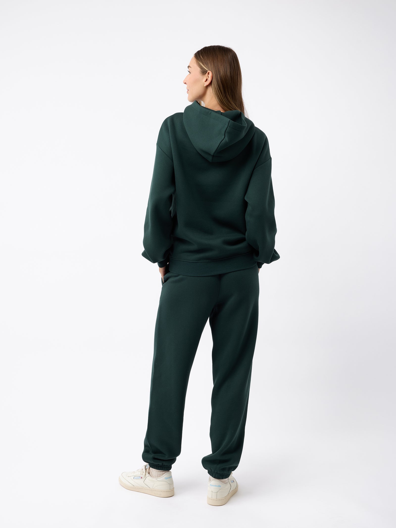 A person stands facing away, wearing a dark green Women's CityScape Hoodie by Cozy Earth with matching sweatpants. They have long hair and are also wearing white sneakers. The background is plain white. 