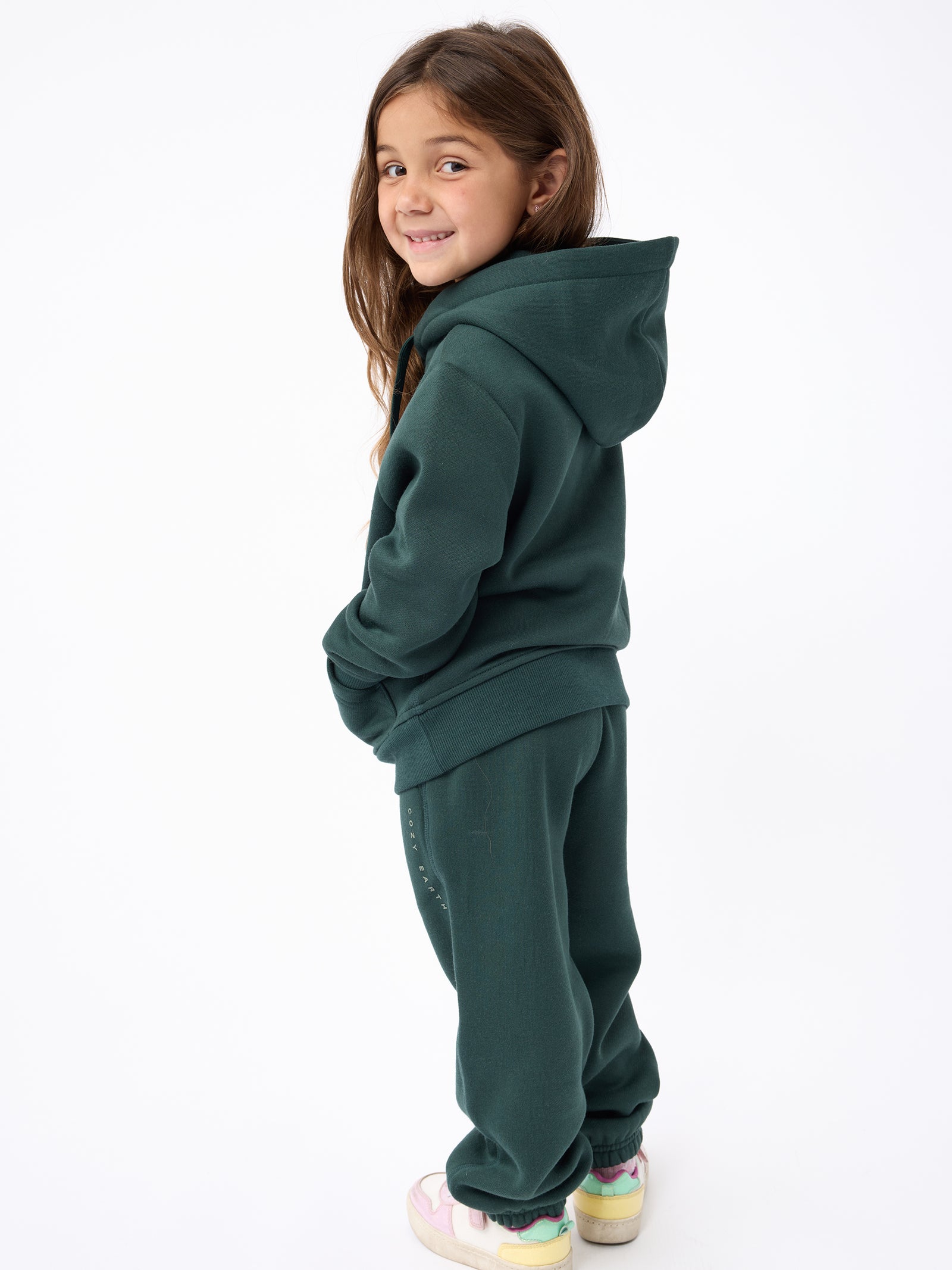 A young girl with long brown hair is smiling and standing sideways, wearing a Cozy Earth's dark green Kid's CityScape Hoodie and matching sweatpants. She has on pink and yellow sneakers, set against a plain white background. 