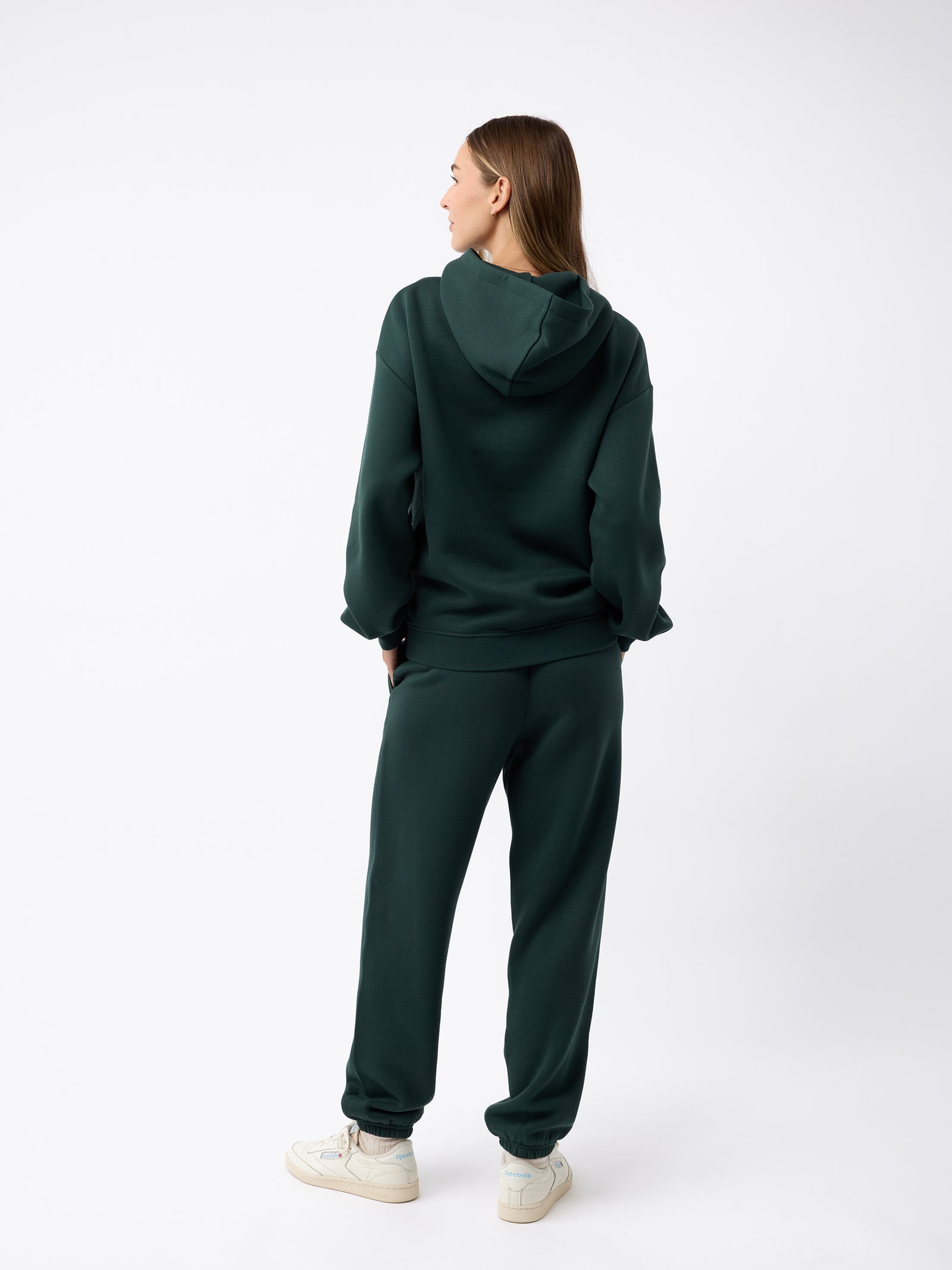 A person with long hair is wearing Cozy Earth's Women's CityScape Sweatpant in dark green, paired with a matching hoodie, and standing against a plain white background. They are turned away, gazing slightly to the side, showcasing a casual and relaxed style. 