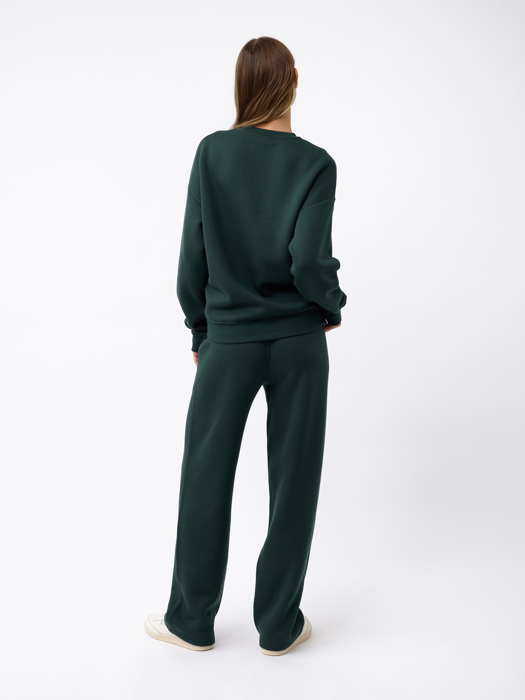 A person stands facing away, dressed in a dark green Cozy Earth sweatshirt and Women's CityScape Wide Leg Pant, with their hands tucked into the pockets. The individual has long brown hair and is wearing white sneakers. The background is plain white. |Color:Spruce