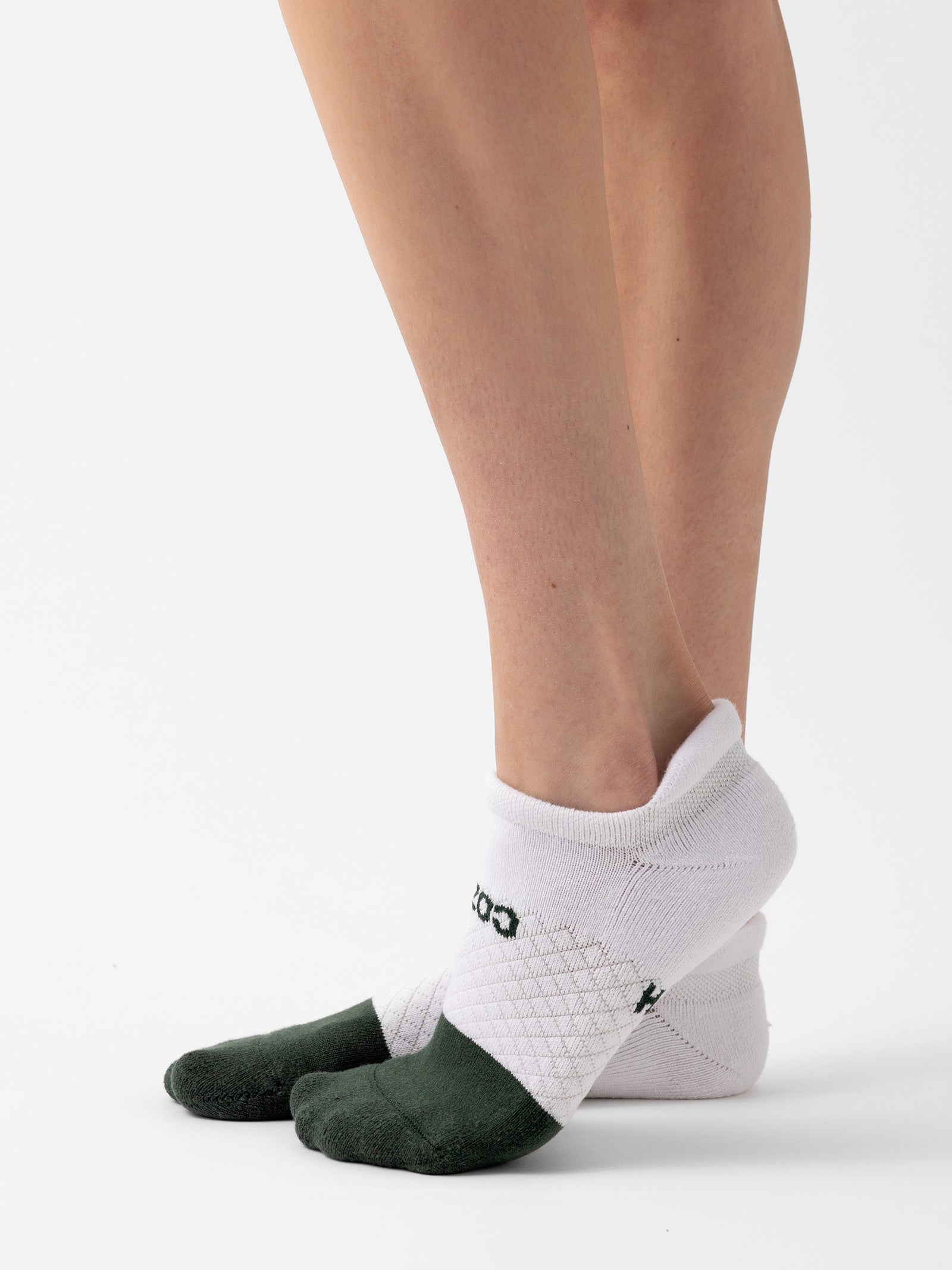 A person's lower legs and feet are shown wearing white socks with green toe and heel sections from the Cozy Earth Essential Ankle Sock 2-Pack. The letters "COZ" are visible on the side of one sock. The background is a plain white surface. 