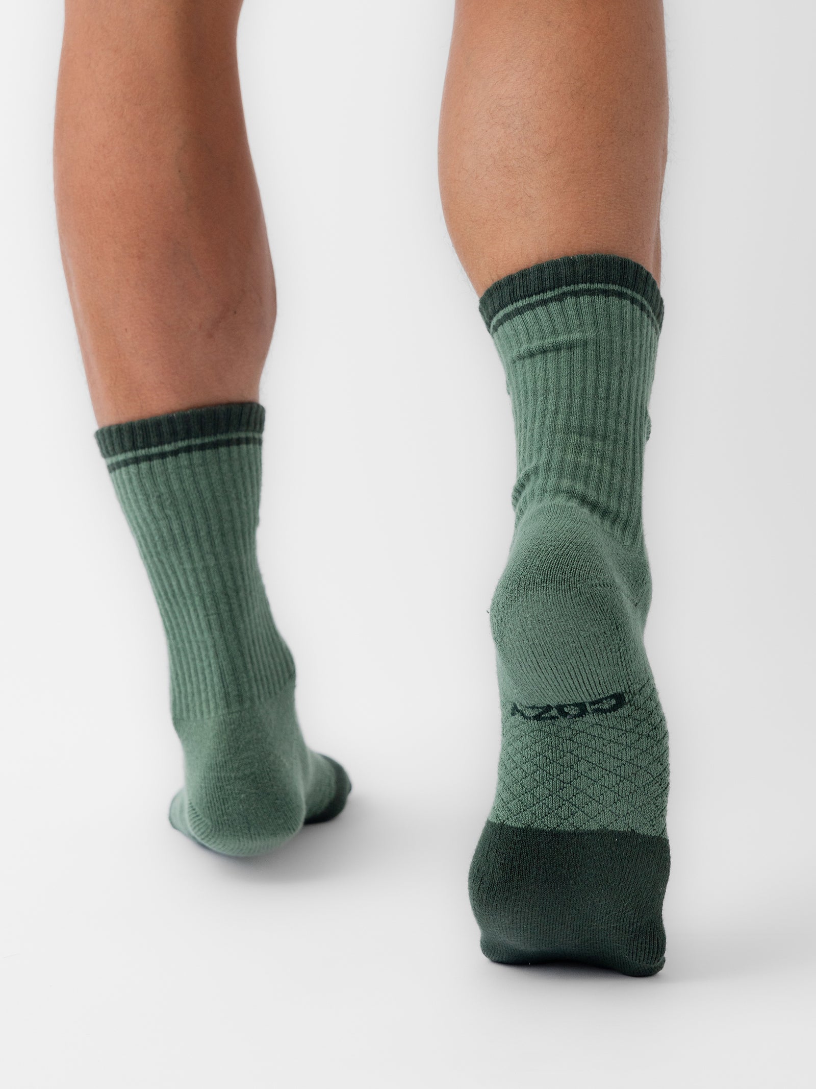 Close-up view of an individual's lower legs and feet, showcasing them wearing green, ribbed crew socks from the Essential Calf Sock 2-Pack by Cozy Earth. The socks have a dark green band at the top and heel, feature honeycomb-like cushioning at the bottom, and display text on the left sock. The background is white. 