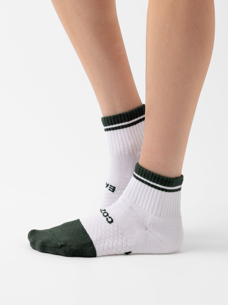 A person wearing Cozy Earth's Essential Quarter Sock, which features a white base with dark green toes, heels, and cuffs. The socks also have dark green stripes near the cuffs and text on the sides. The background is plain white.