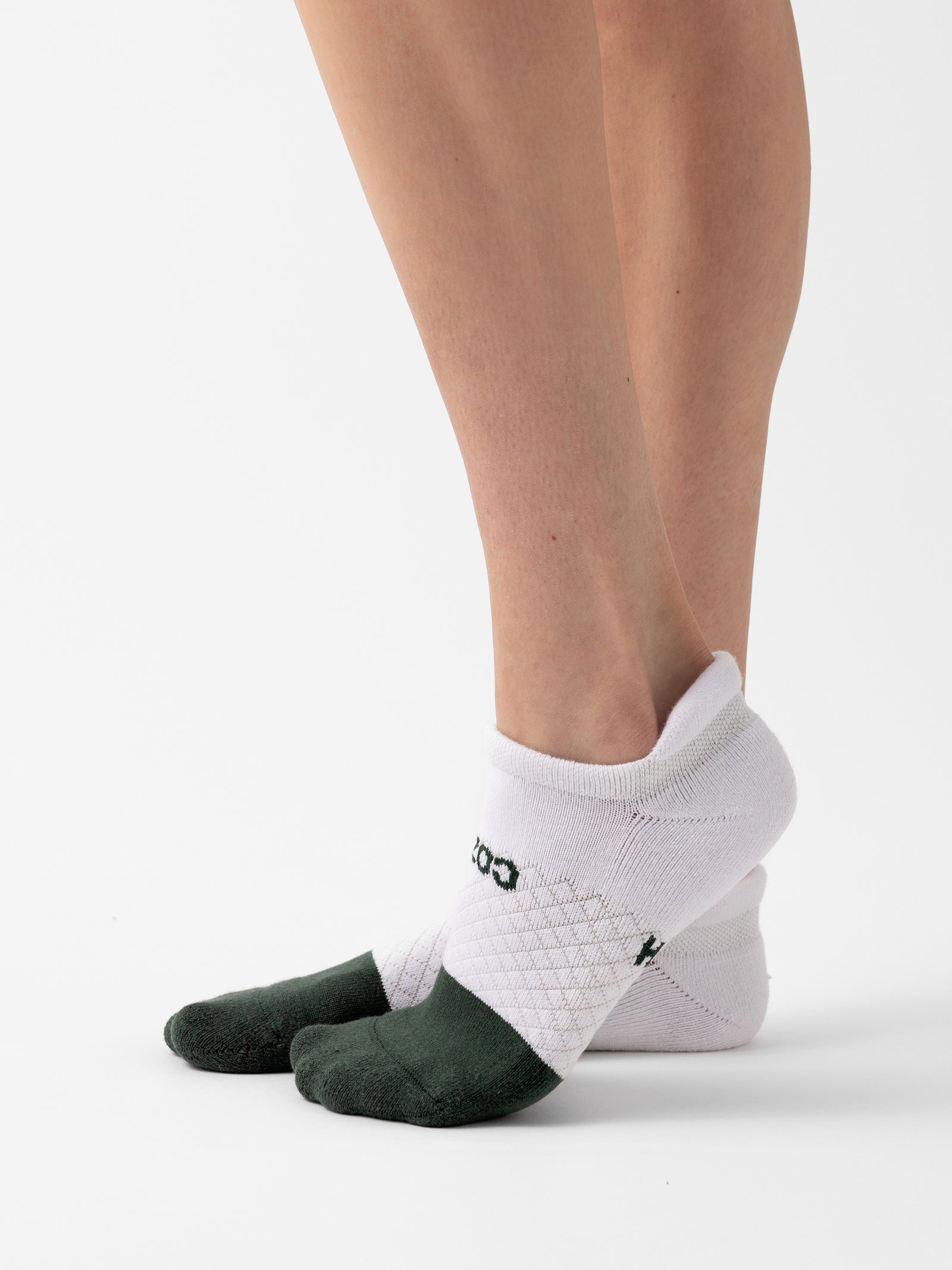 Close-up view of a person's legs and feet wearing white Cozy Earth Essential Ankle Socks with green accents on the toes, standing on a white background. The socks have the word "GOOD" written in black text near the ankle area. 