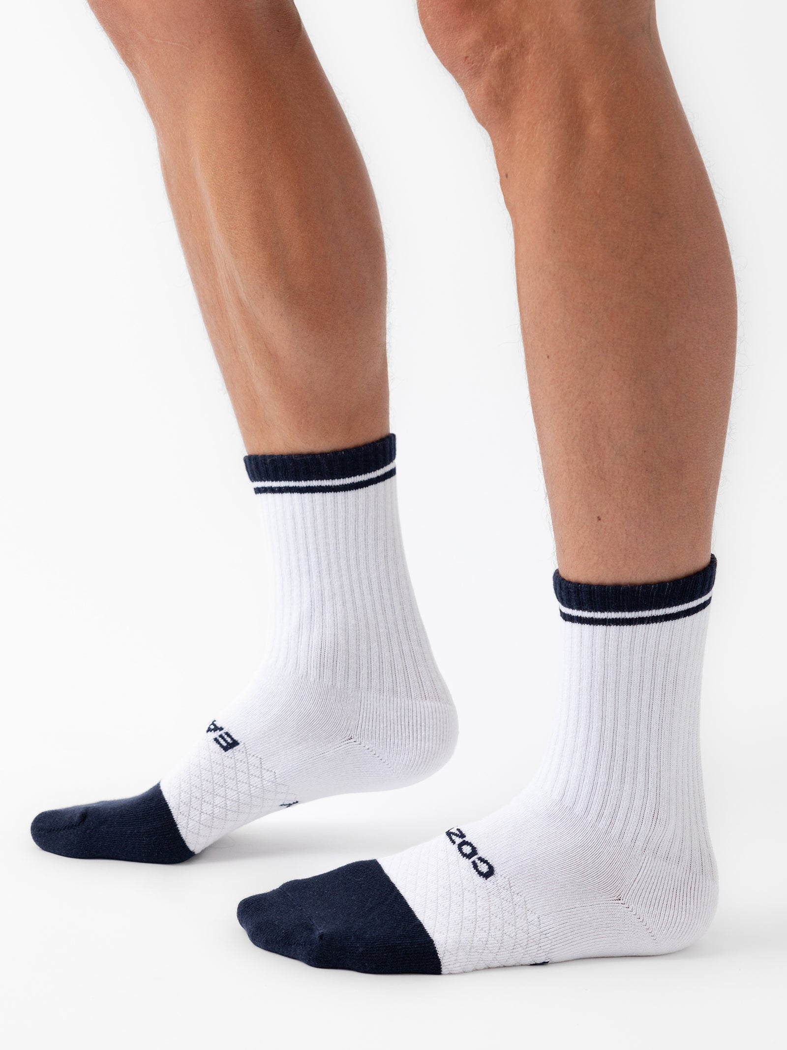 Close-up of a person's lower legs and feet wearing Cozy Earth's white Essential Calf Sock with black toe and heel accents. The socks feature black stripes around the top, and the person is standing on a white background. 