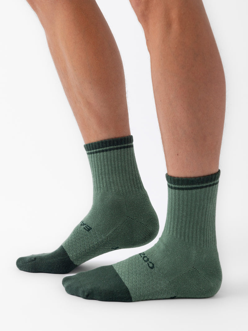 Two legs are adorned with a pair of Cozy Earth's Essential Quarter Socks from their 4-Pack collection, featuring a mid-calf length in green with darker green accents on the toes and heels. The socks exhibit a ribbed texture around the ankle and a knit pattern on the foot, as the person stands against a white background.