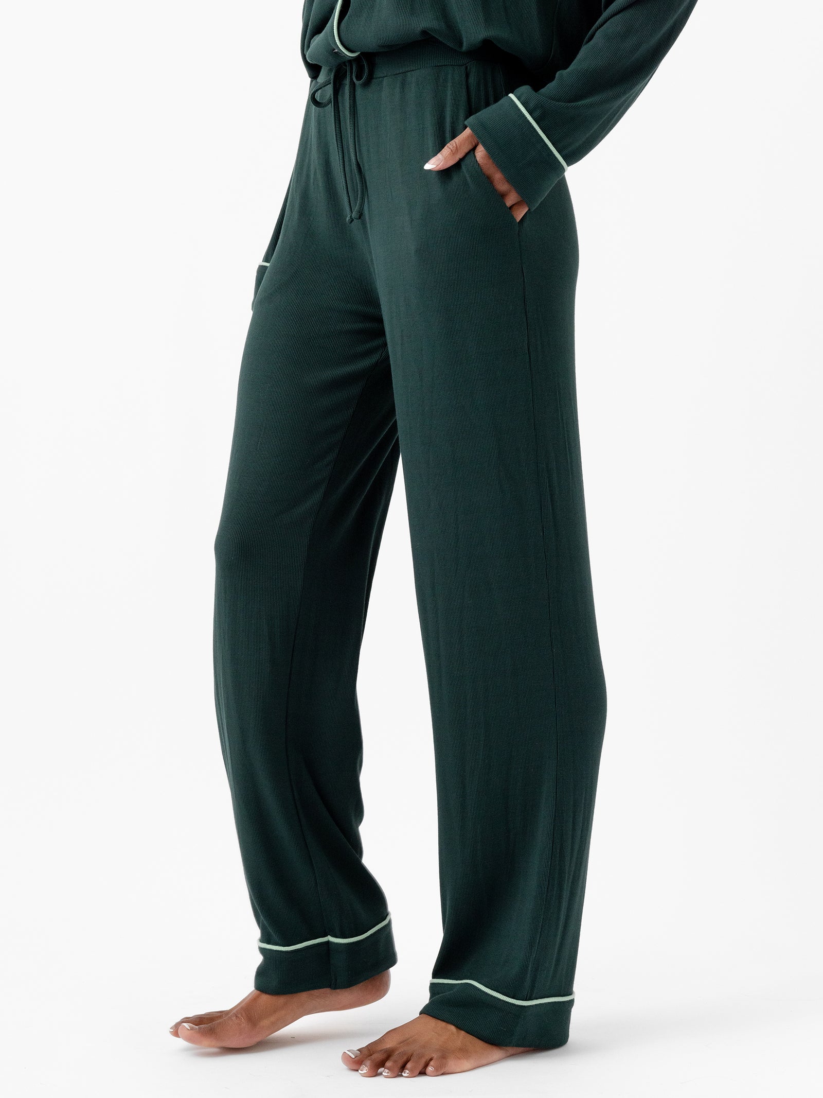 A person wearing Cozy Earth's Women's Bamboo Rib Knit Classic Pajama Pants in dark green, featuring a drawstring waist and side pockets, is standing barefoot. The pants have a relaxed fit and are accented with light green piping at the cuffs. The person's hands are in their pockets against a plain white background. 