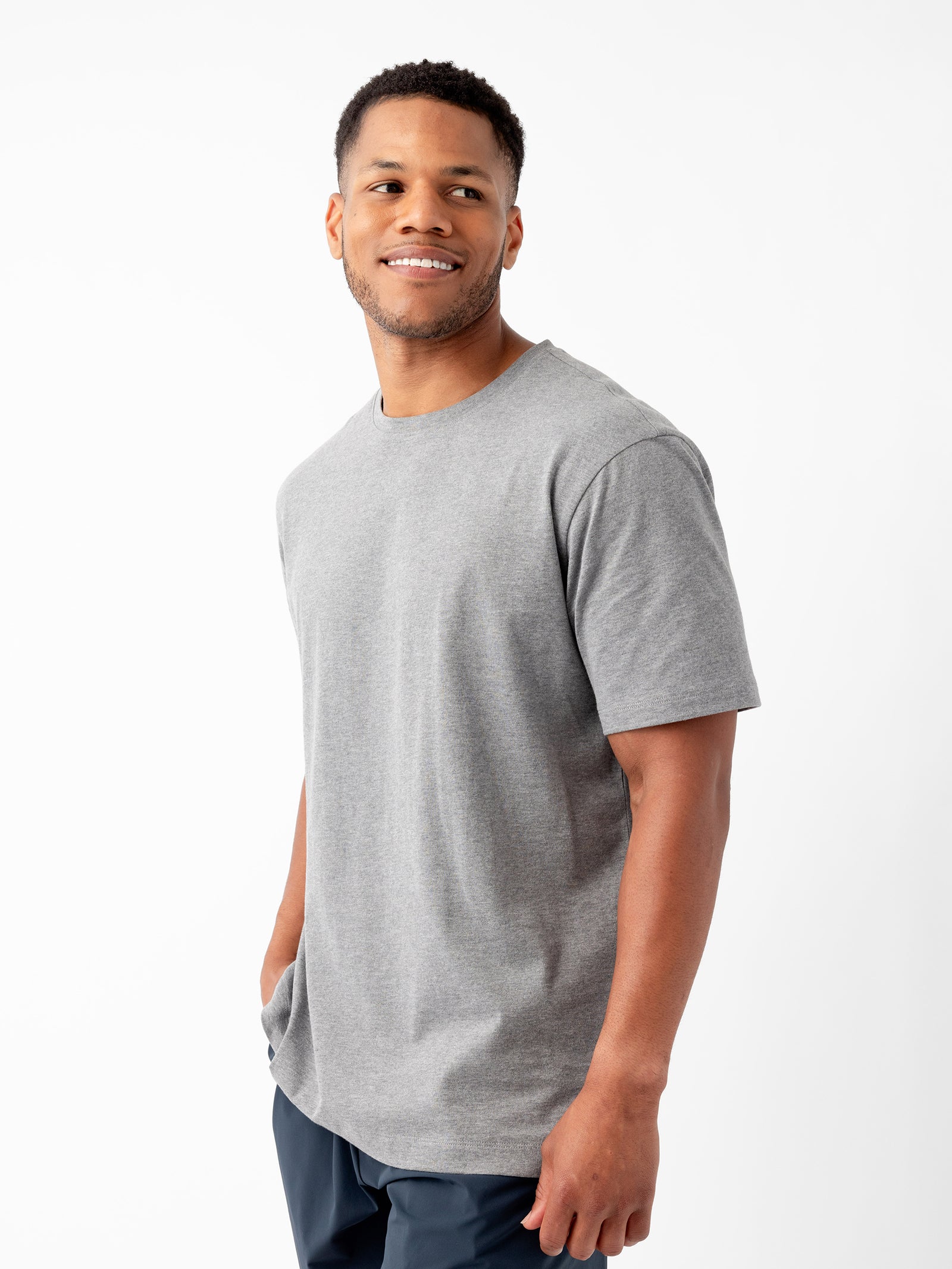 A man with short hair is standing against a white background, wearing a grey Men's All Day Tee from Cozy Earth and dark pants. He is smiling and looking slightly to his right, with one hand in his pocket. 