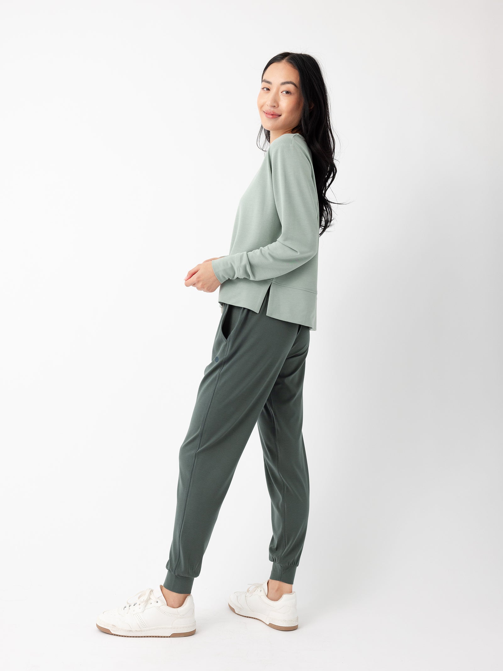 A woman with long dark hair is standing sideways, looking over her shoulder and smiling. She is dressed in a light green long-sleeve shirt, Cozy Earth's dark green Women's Studio Jogger, and white sneakers. The background is plain white. 