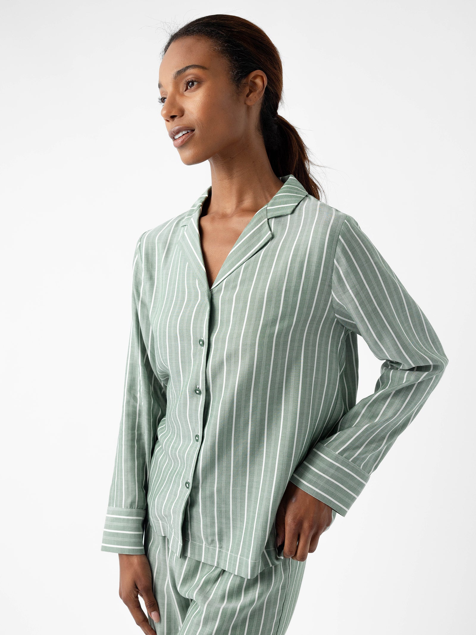 A woman dressed in the Cozy Earth's Women's Soft Woven Long Sleeve Pajama Top, featuring a green and white striped design, stands against a simple white backdrop. Her hair is tied back as she gazes to the side with a relaxed and thoughtful expression. 