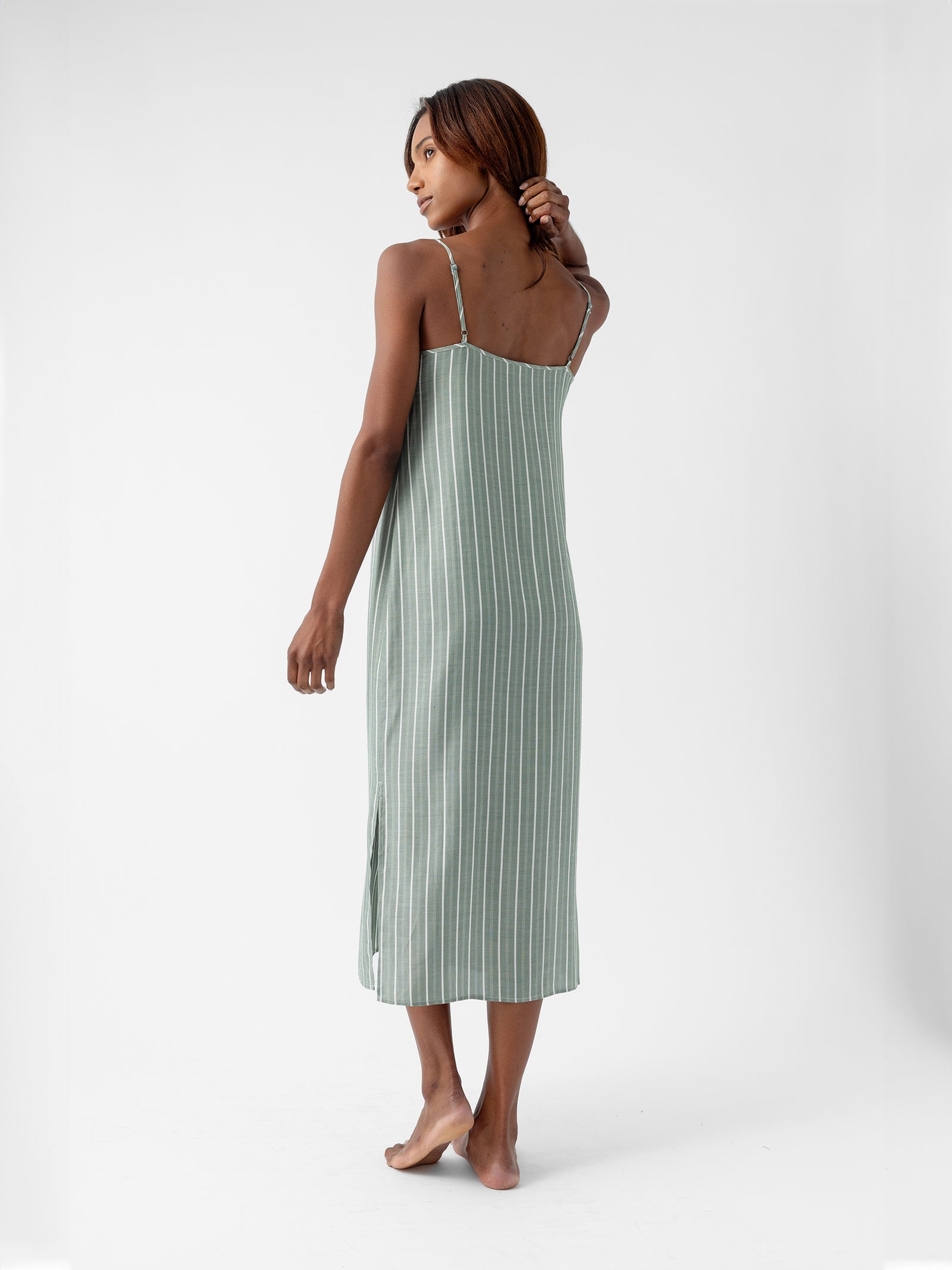 A woman stands barefoot in a long, green, vertically striped Women's Soft Woven Nightgown by Cozy Earth. She slightly turns her head to the left with one hand touching her head, set against a plain white background. 