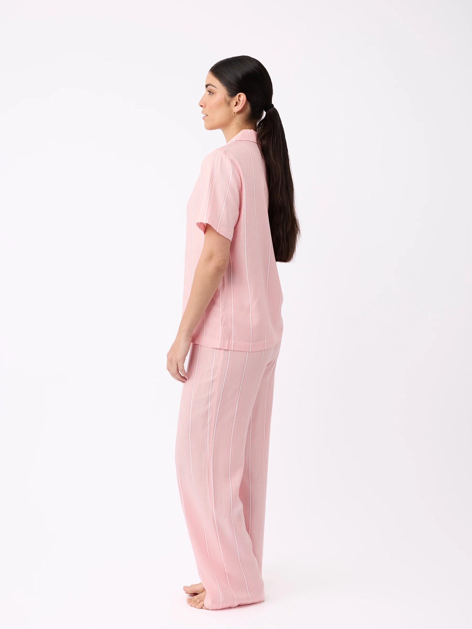 A woman with long, dark hair tied in a ponytail stands barefoot and sideways in a white space, wearing Cozy Earth's pink short-sleeved shirt and Women's Soft Woven Pajama Pant. 