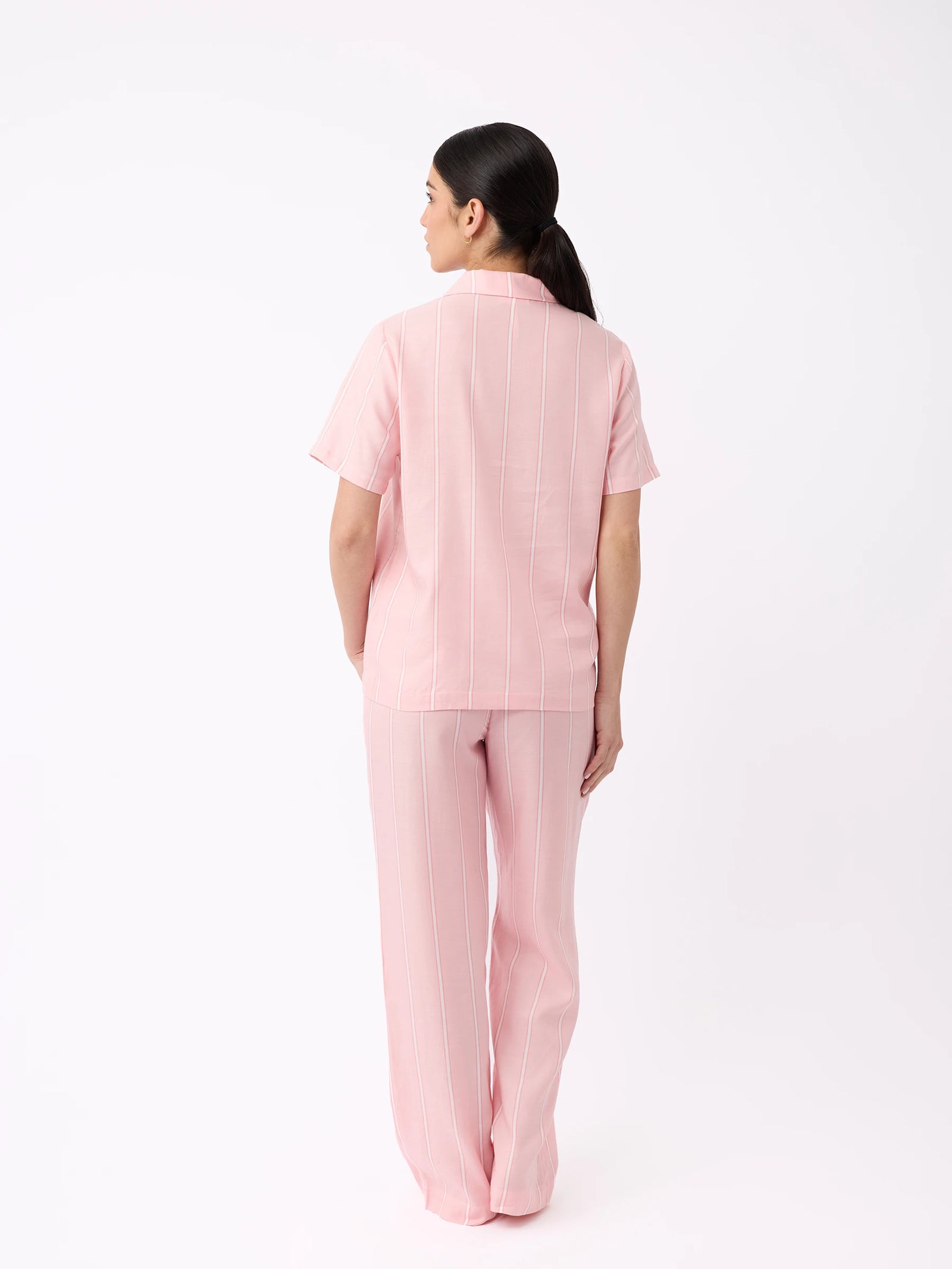 A person with long dark hair stands with their back to the camera, wearing a light pink outfit resembling Cozy Earth's Women's Soft Woven Short Sleeve Pajama Set against a plain white background. 