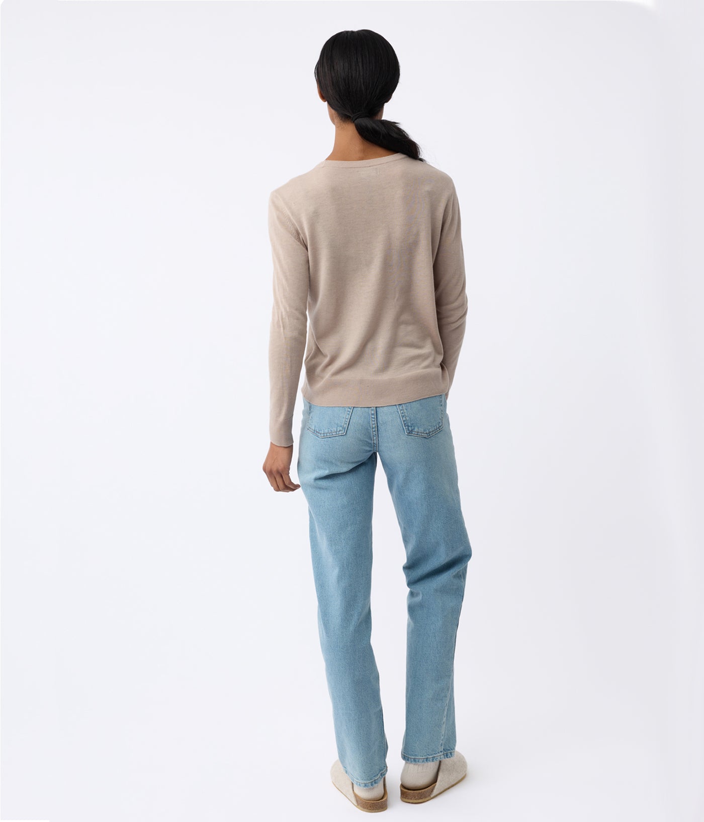 A woman with long hair, facing away, wears a Cozy Earth Women's AirKnit Crewneck Sweater in beige, light blue jeans, and beige shoes against a plain white background. Color:Taupe