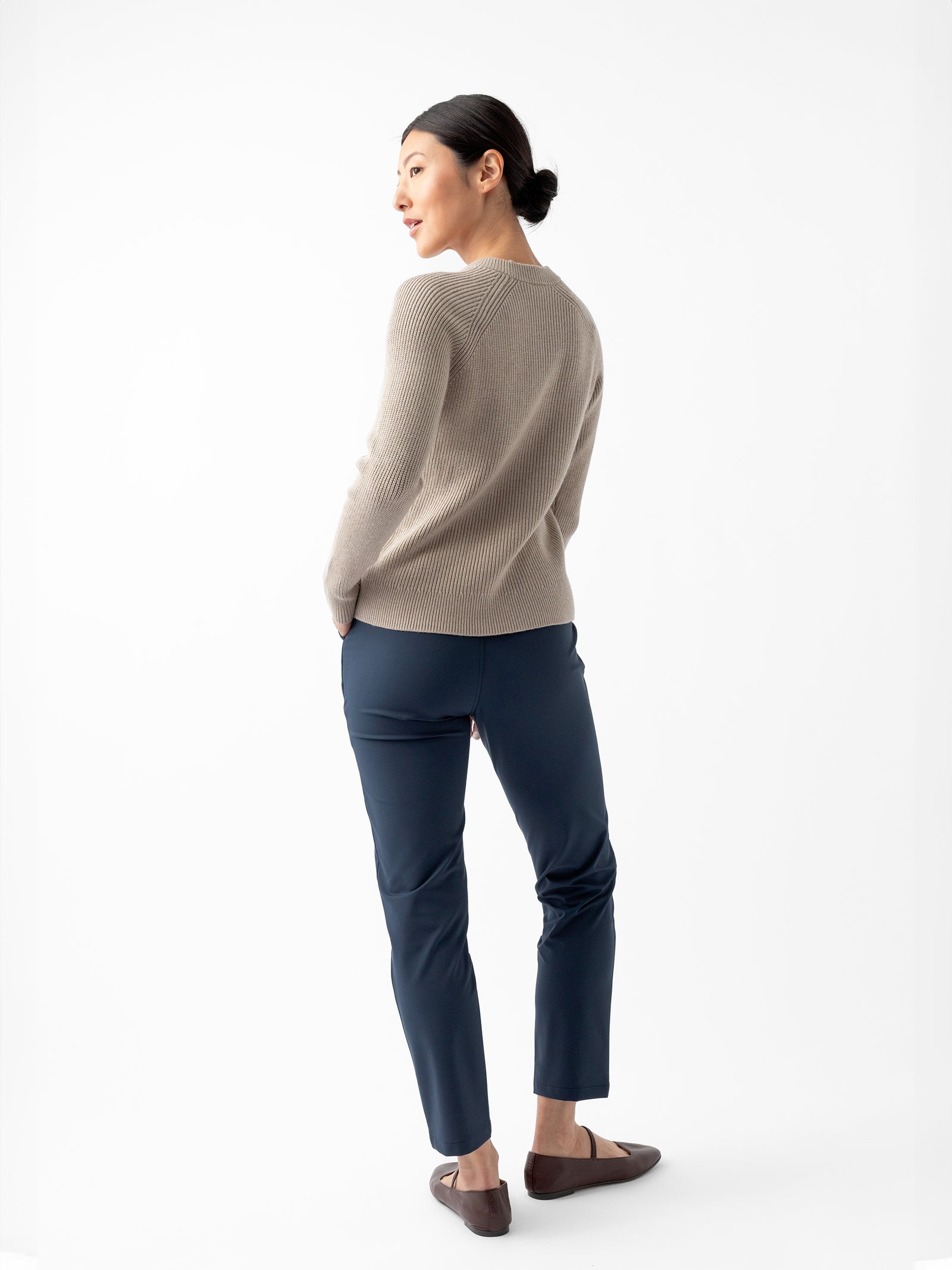 A person is standing against a white background, facing slightly to the side. They are wearing Cozy Earth's Women's Classic Crewneck in beige, dark blue pants, and brown shoes. Their hands are tucked into their pants' pockets, and their dark hair is pulled back in a low bun. 