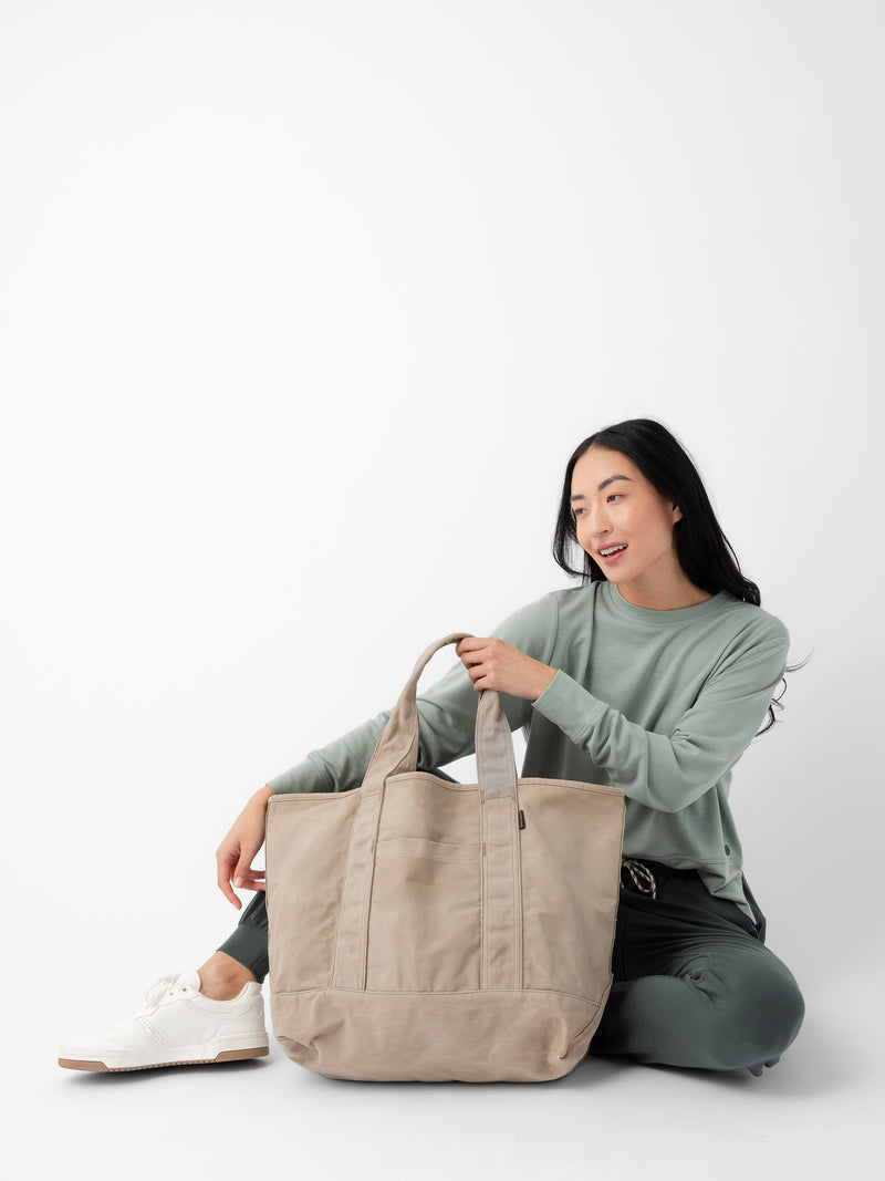 A person with long dark hair, dressed in a green long-sleeve shirt and dark pants, sits on the floor against a white background. They are holding a large Cozy Earth Waxed Canvas Tote and appear to be smiling, with white shoes visible beside them.