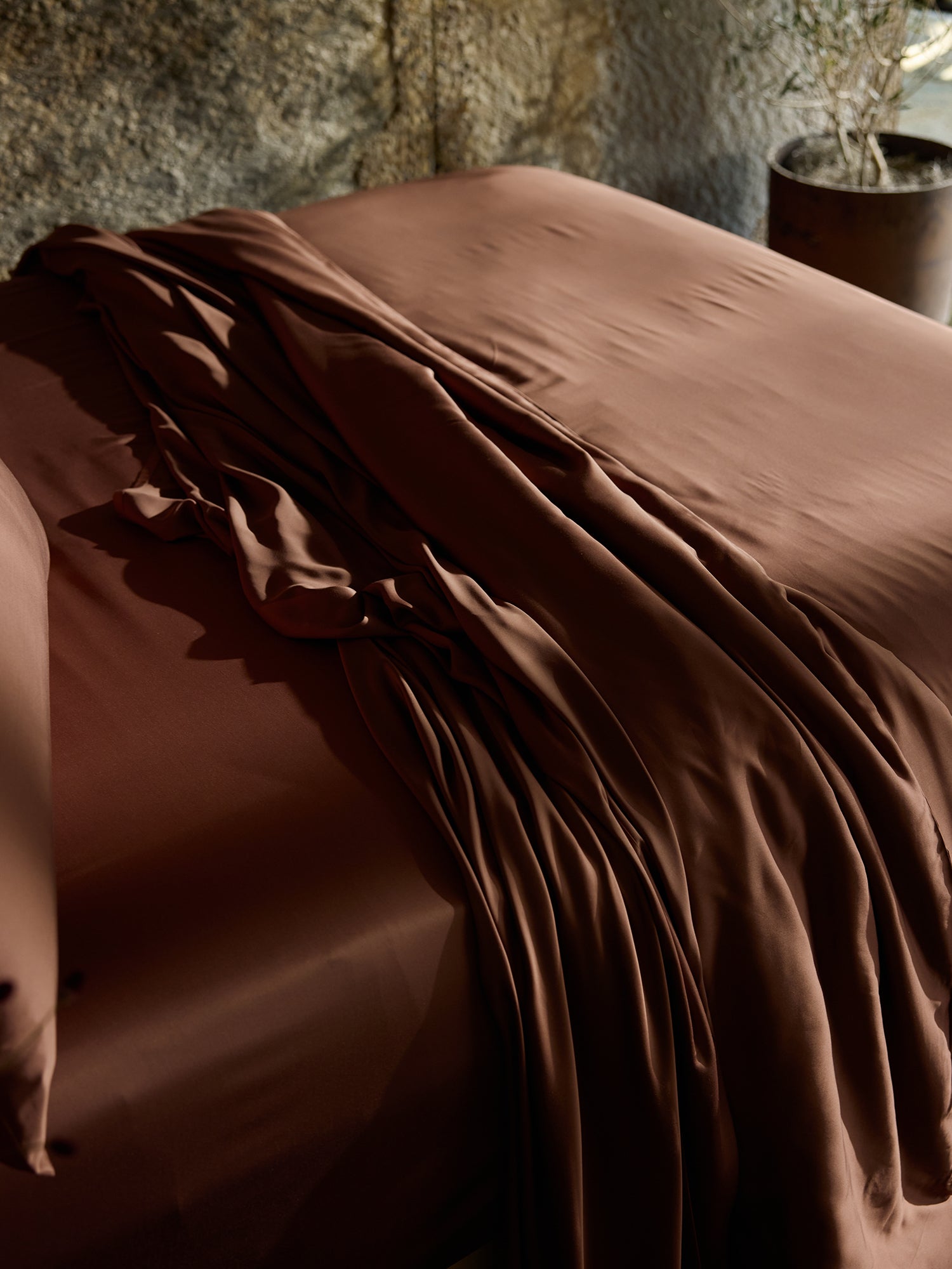 A bed adorned with a luxurious Cozy Earth Bamboo Duvet Cover in rich brown, complete with matching pillowcases, is bathed in gentle natural light. A long draped fabric, possibly another duvet or extra sheet, cascades over the bed. In the background, a textured wall and a potted plant are visible. 