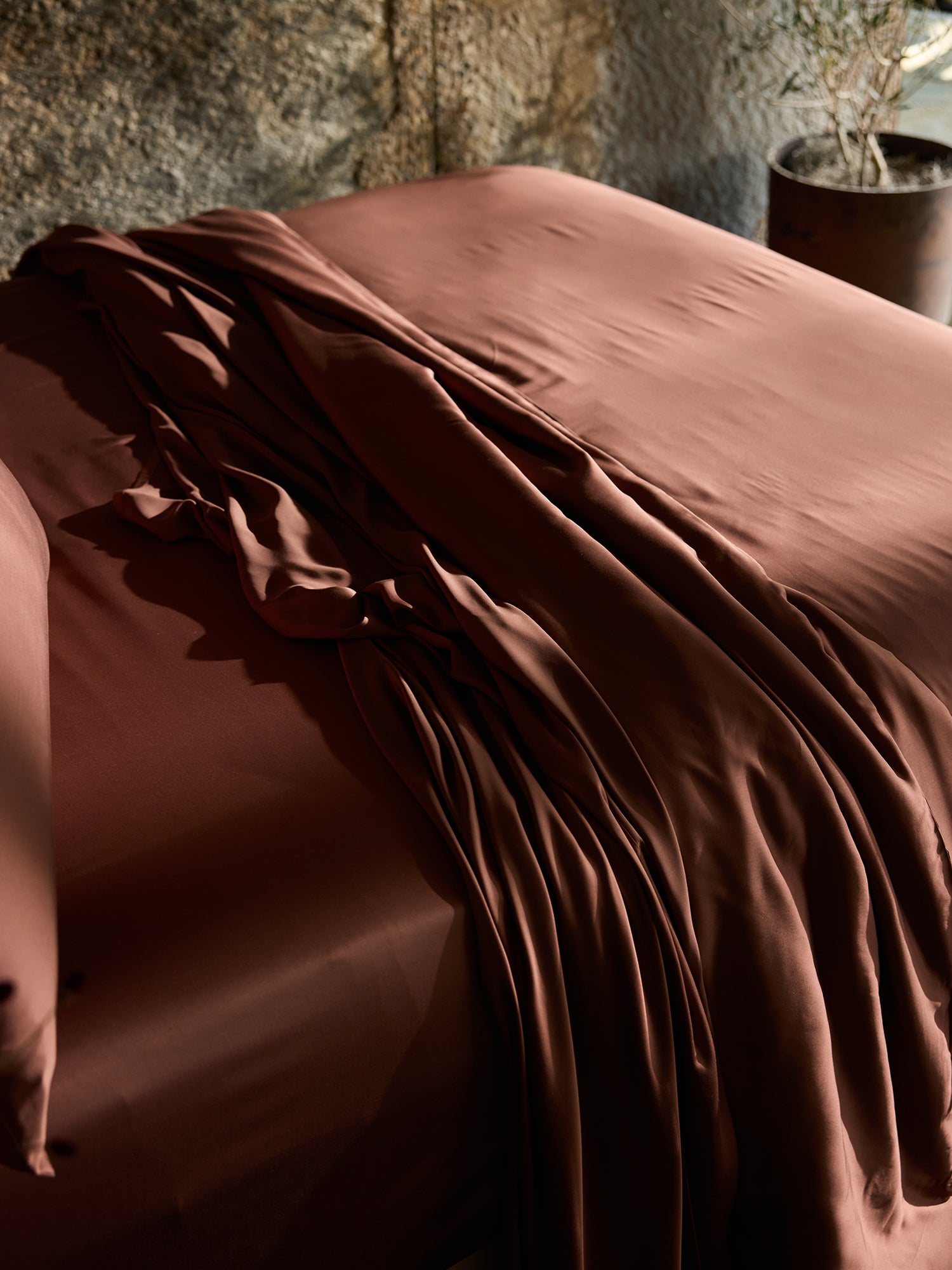 A neatly made bed adorned with Cozy Earth's Bamboo Fitted Sheet in a rich dark reddish-brown hue, creating a luxurious and cozy feel. The background features a textured wall and a potted plant, adding a touch of nature to the setting. The lighting casts a warm and inviting glow on the scene. 