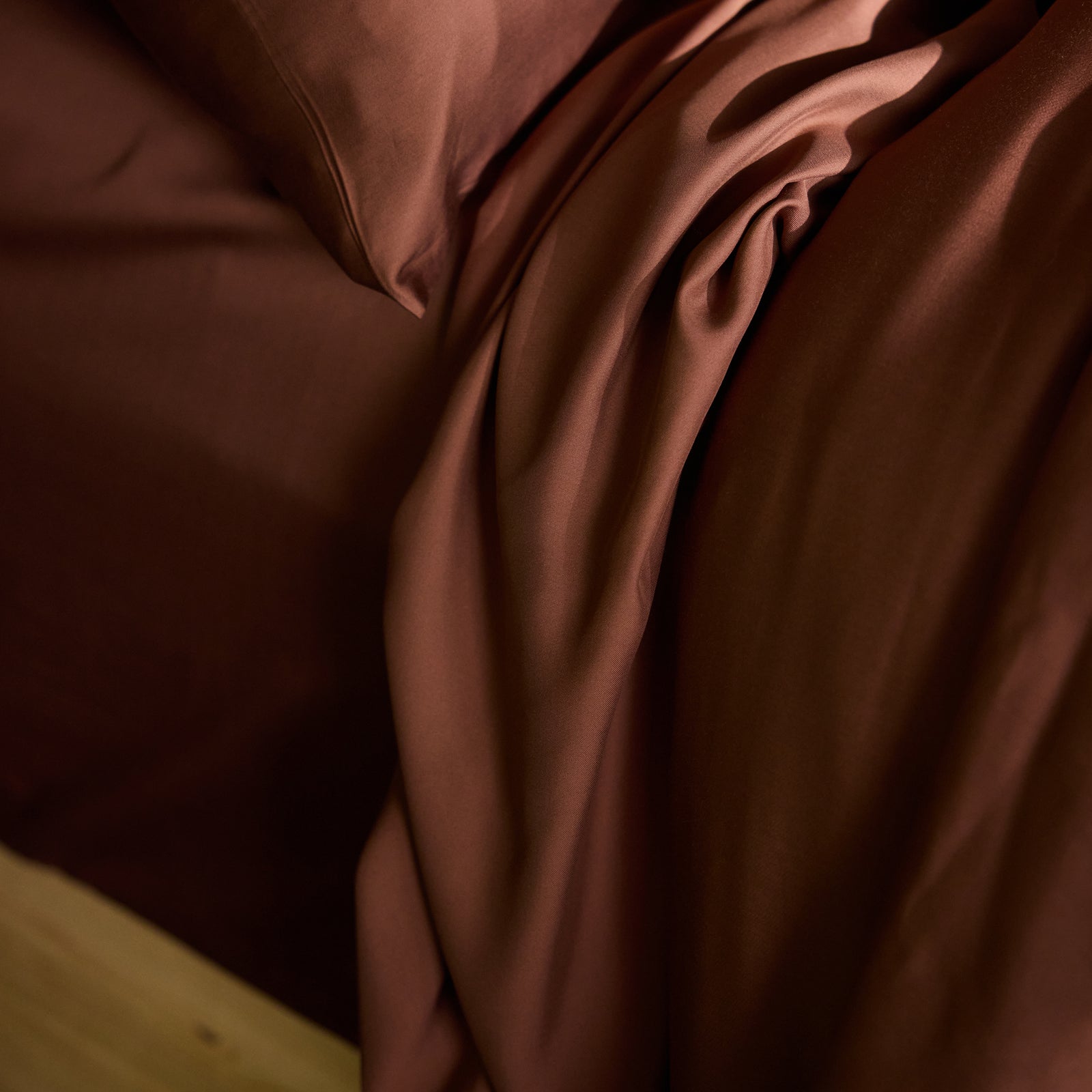 Close-up of a bed featuring Cozy Earth's Bamboo Flat Sheet in deep brown, with a slightly wrinkled, rich brown comforter cascading over the edge. The fabrics have a smooth, silky texture and exude a sense of warmth and coziness. 