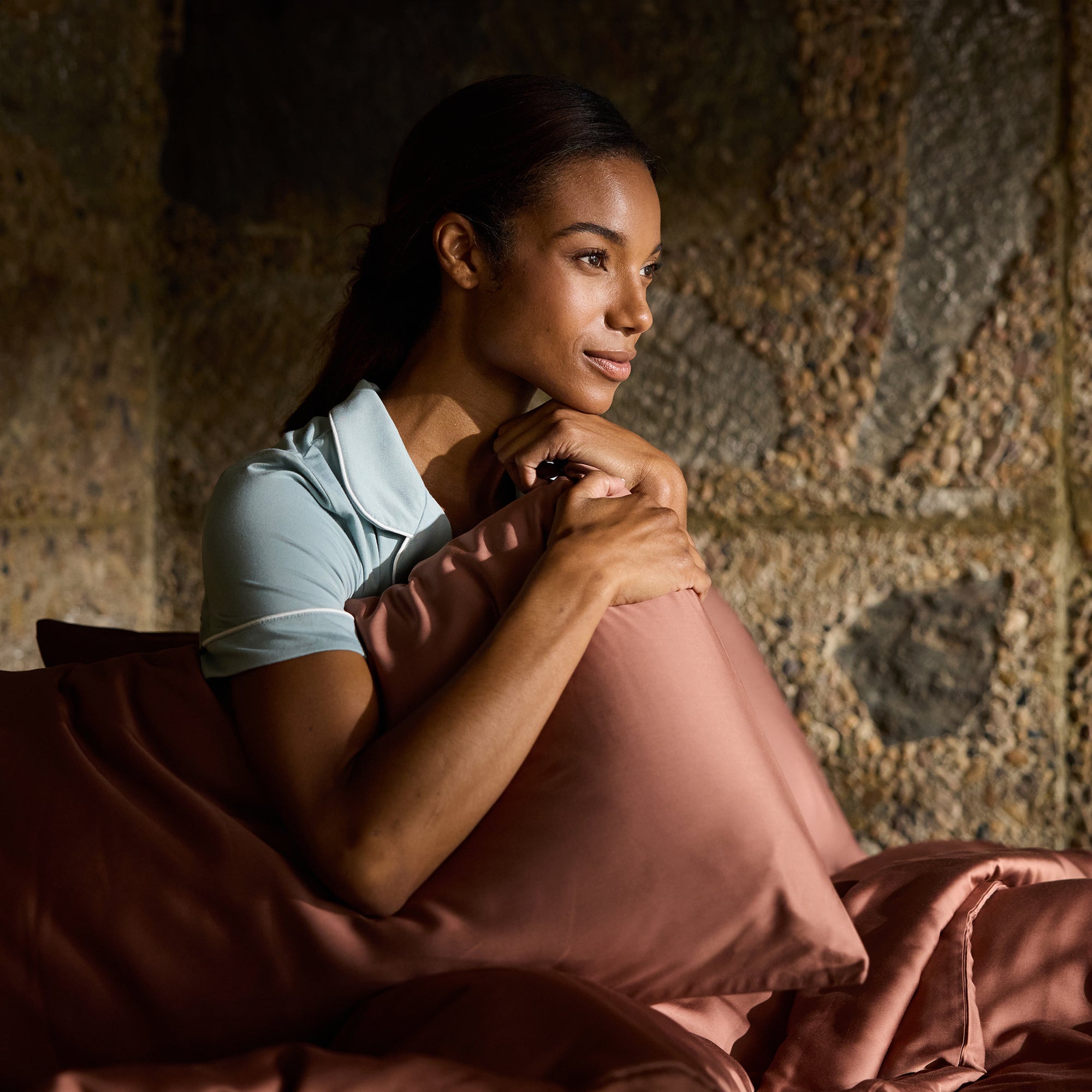 A woman with a serene expression sits in a dimly lit room, holding a pillow close to her chest. She is wearing a light blue shirt and is surrounded by warm, brown-toned bedding from the Bamboo Sheet Set by Cozy Earth. The background features a textured stone wall. 
