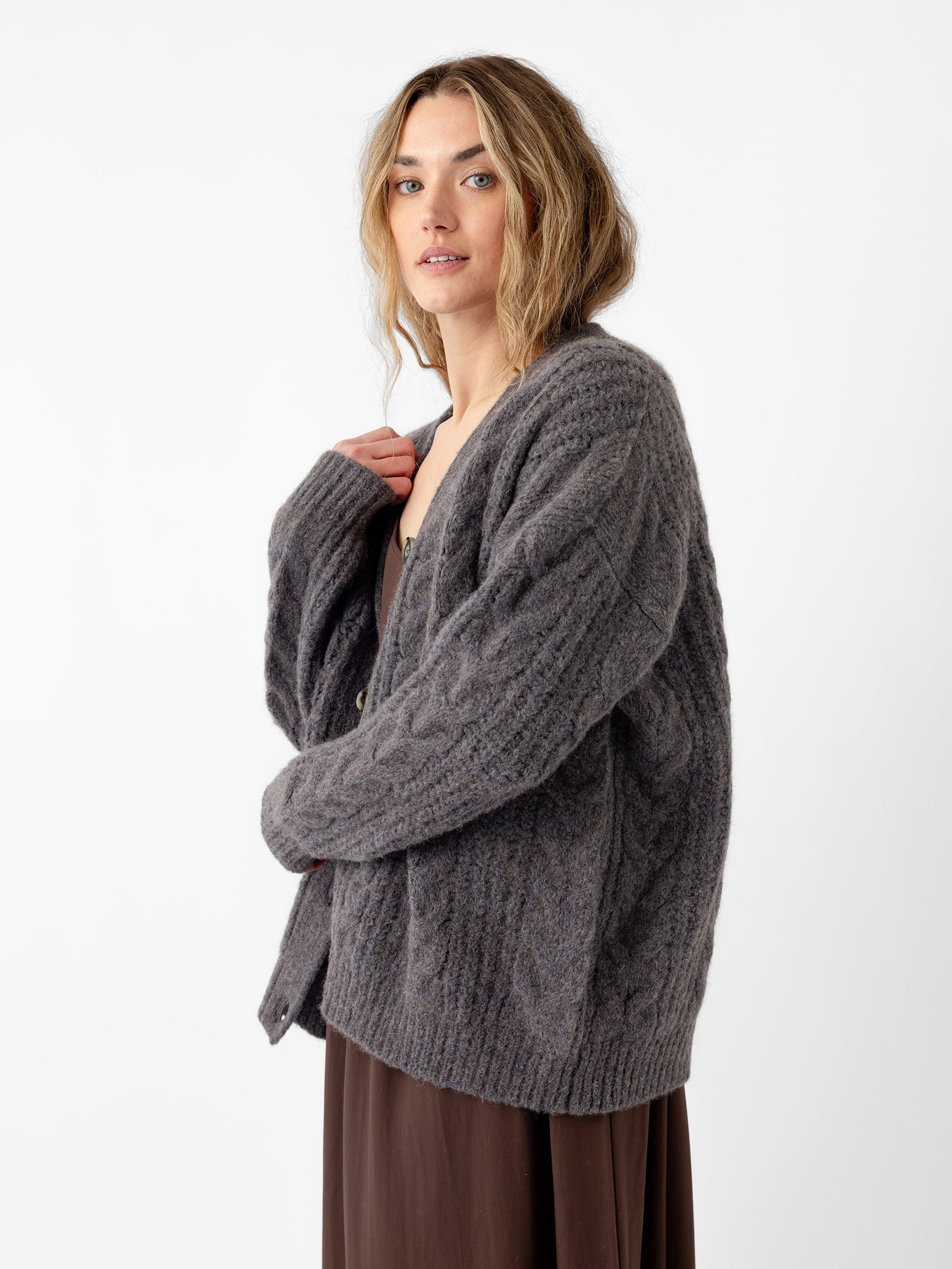 A person with long hair stands against a white background, wearing Cozy Earth's Oversized Cable Knit Cardigan in gray over a brown dress. They maintain a neutral expression as they look towards the camera, gently holding the cardigan collar with one hand. 