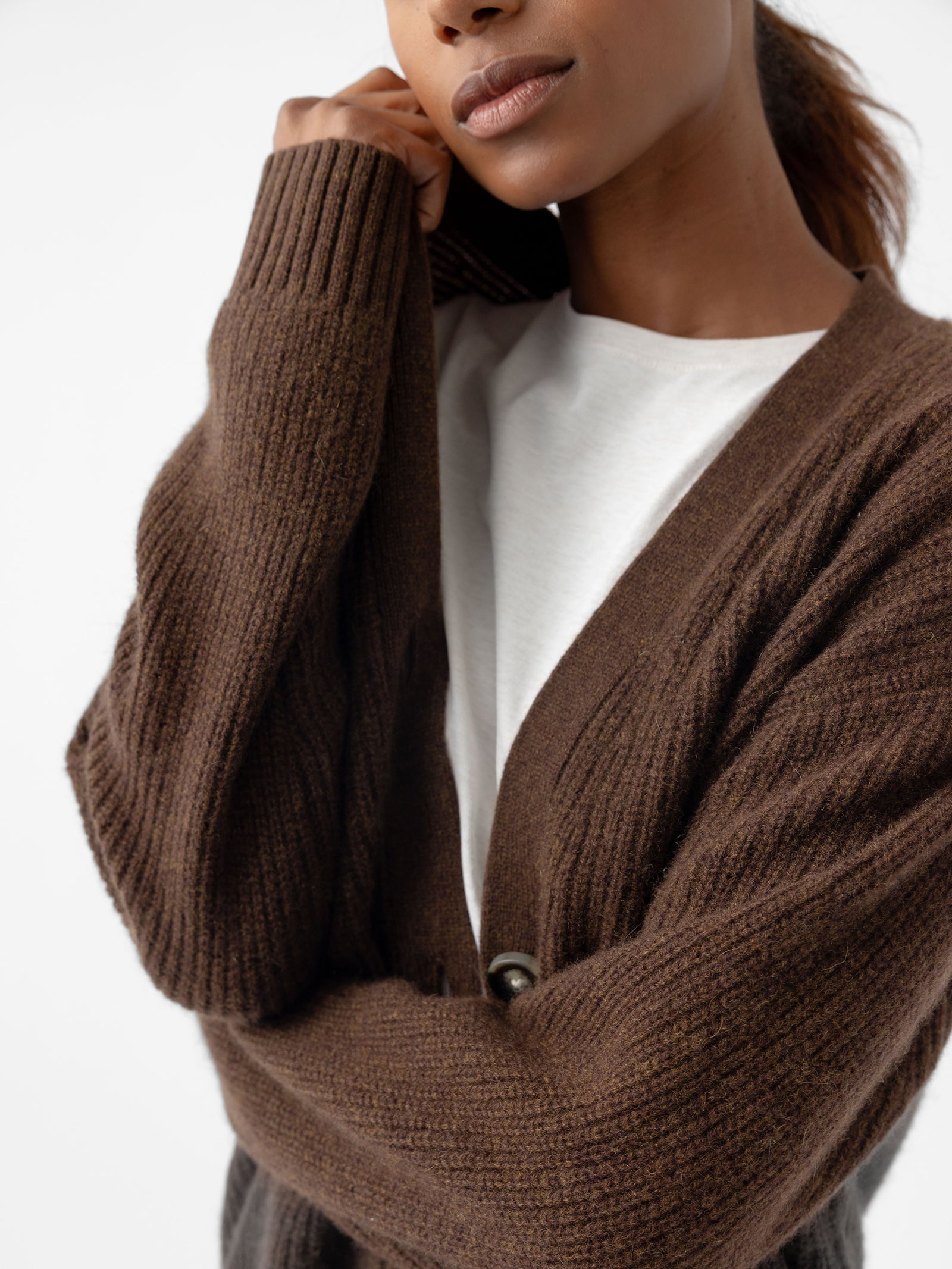 A person is wearing a brown Oversized Sunday Cashmere Cardigan by Cozy Earth over a white top. The cardigan features an oversized fit with large buttons, and the person is softly holding the collar with one hand against a plain white background. 