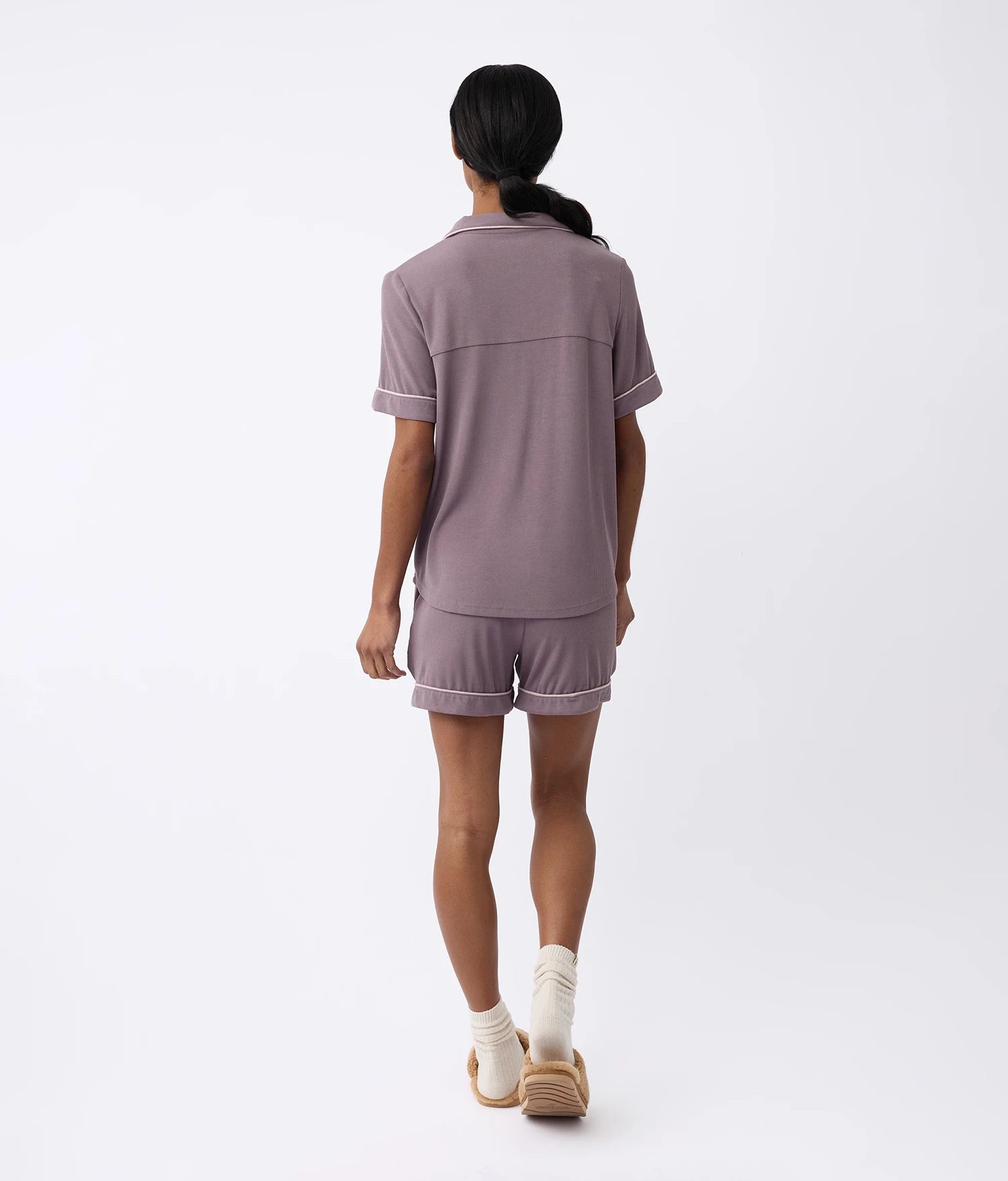 Rear view of a person wearing Cozy Earth's Women's Bamboo Rib-Knit Classic Short Sleeve Pajama Set in purple, paired with white socks and brown slippers, against a plain white background. 