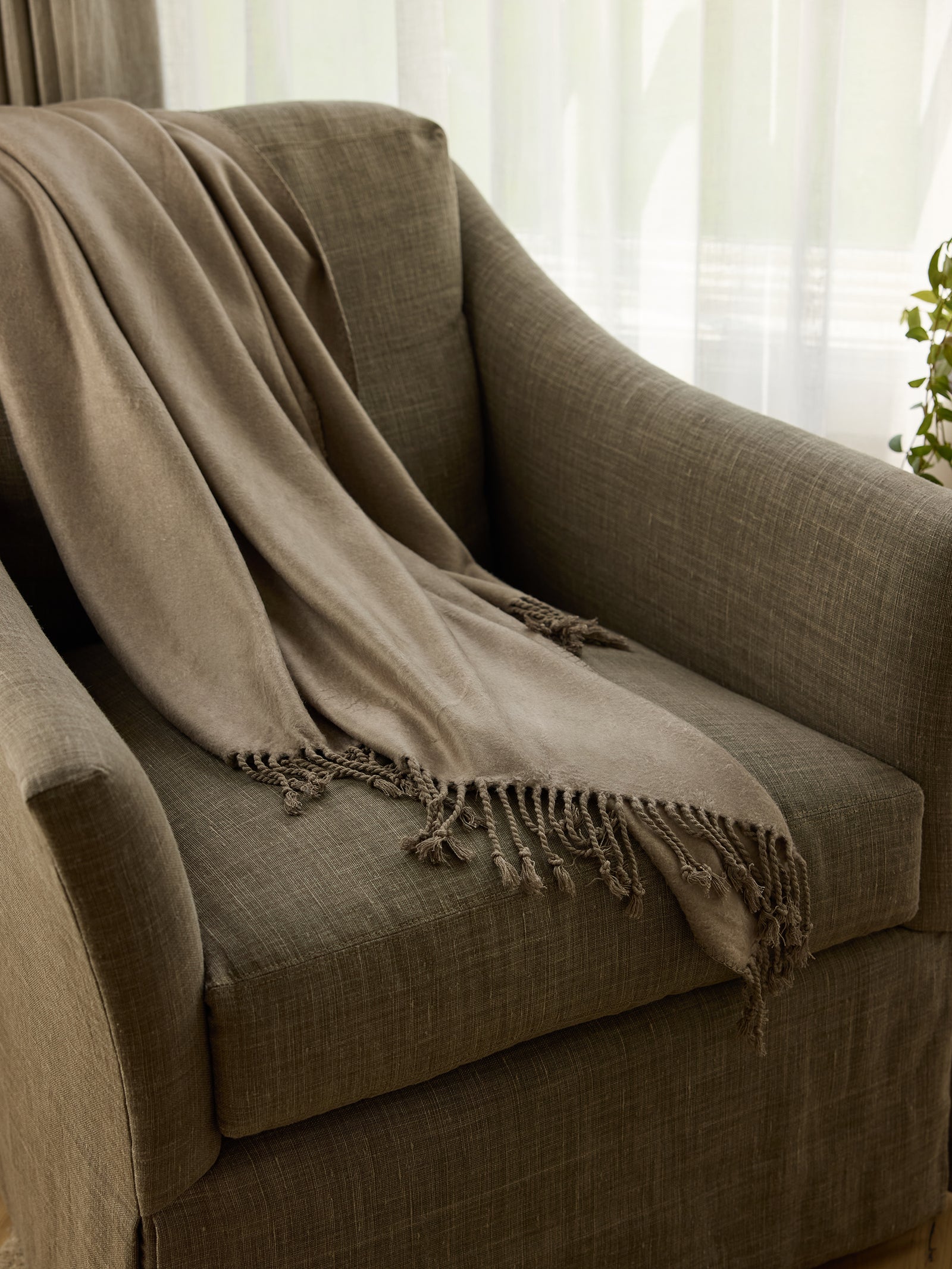 A cozy, olive green armchair with a Bamboo Tassel Throw from Cozy Earth, draped over it. The throw blanket is soft and features fringe detailing at the ends. The chair is positioned in front of white, sheer curtains, with a small leafy plant visible on the side. 