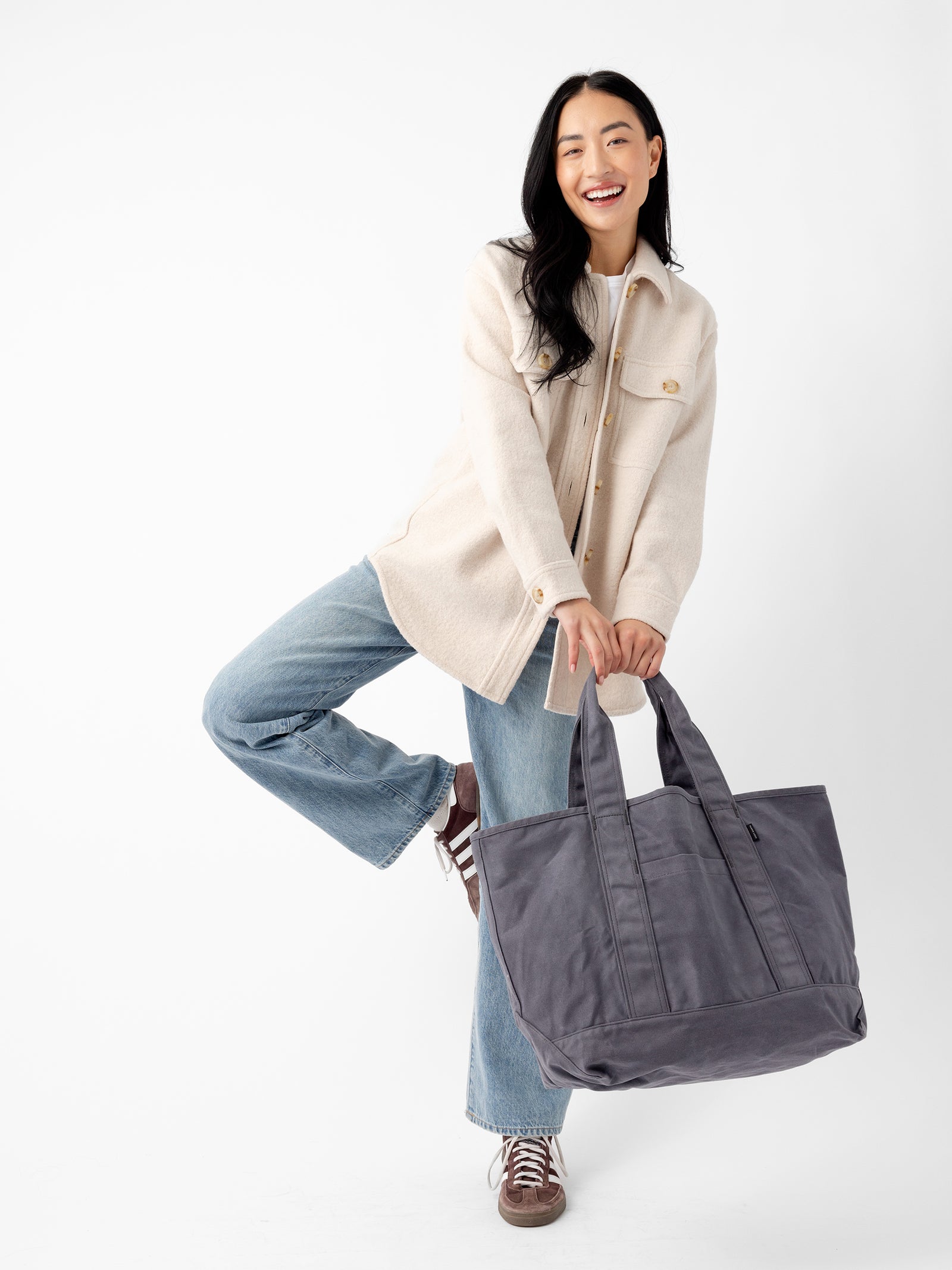 A smiling person with long dark hair is wearing a beige jacket and blue jeans, holding a large Waxed Canvas Tote by Cozy Earth in one hand. They are posing with one leg bent, balancing on the other, and wearing brown sneakers. The background is white. 