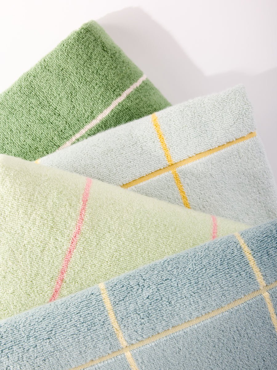 The close-up features four Windowpane Resort Towels by Cozy Earth in soft pastels, showcasing simple grid and line patterns that create a textured look. Shadows add depth, enhancing the clean and minimalistic feel. 