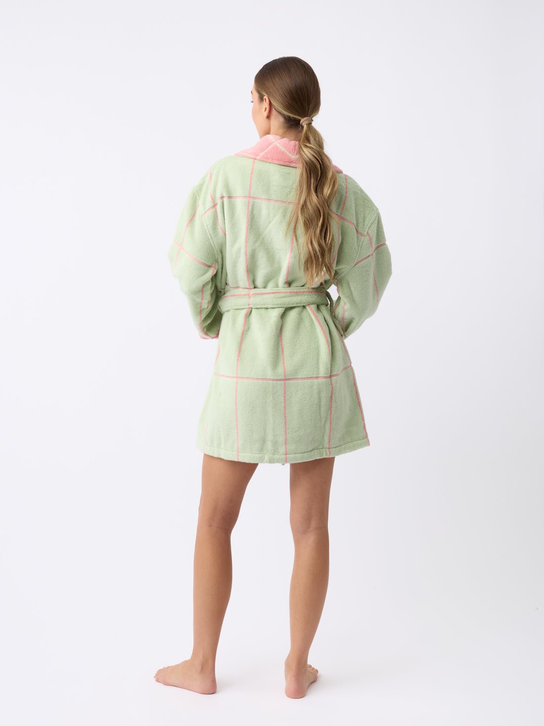 A barefoot individual with long hair stands facing away, wearing the Windowpane Resort Robe in light green and pink checkered design by Cozy Earth against a plain white background. 