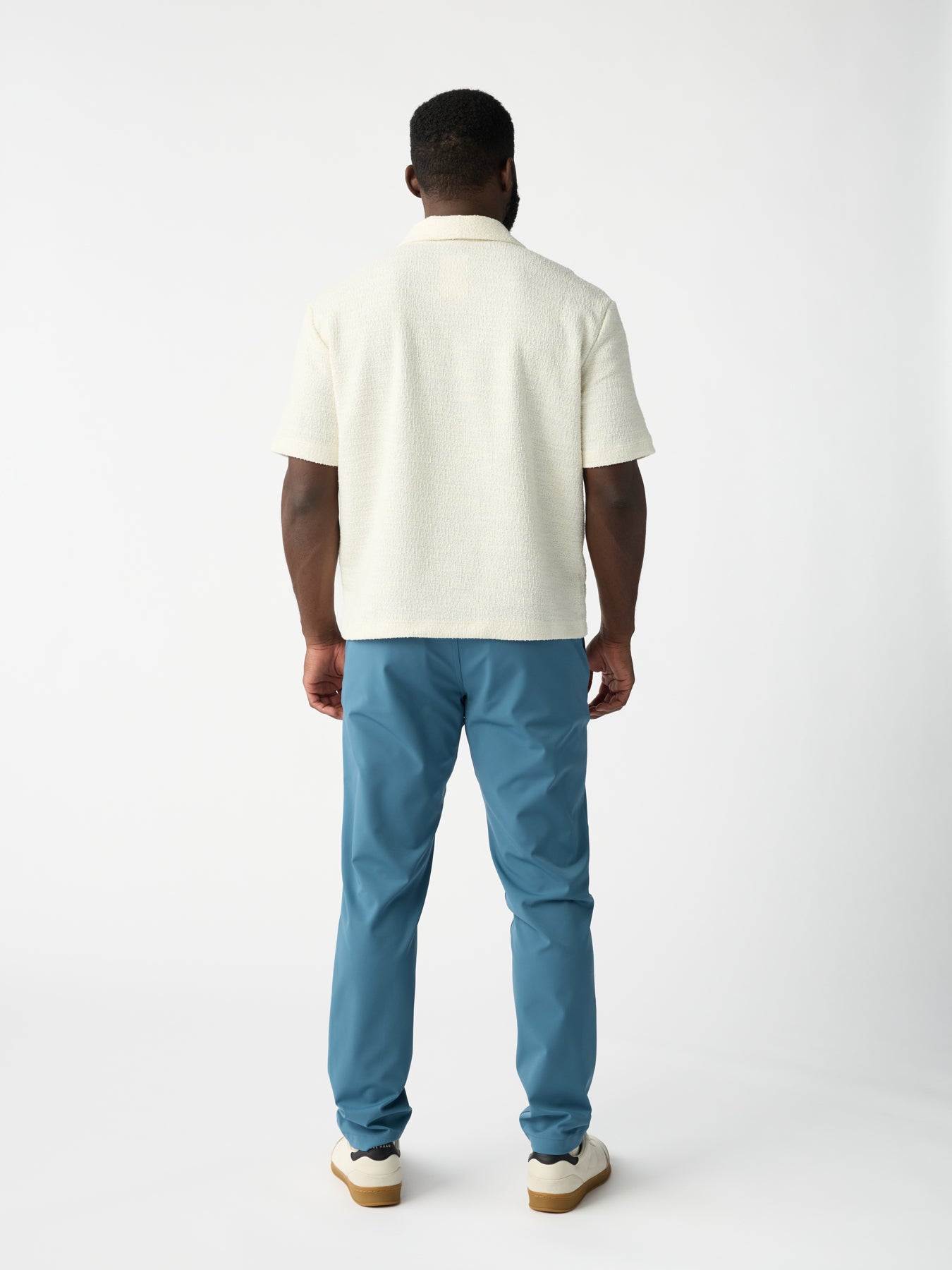 A man with short hair faces away, wearing a textured white shirt and Cozy Earth's light blue Men's Everywhere Pant against a plain white background. His sneakers have white tops with brown soles. 