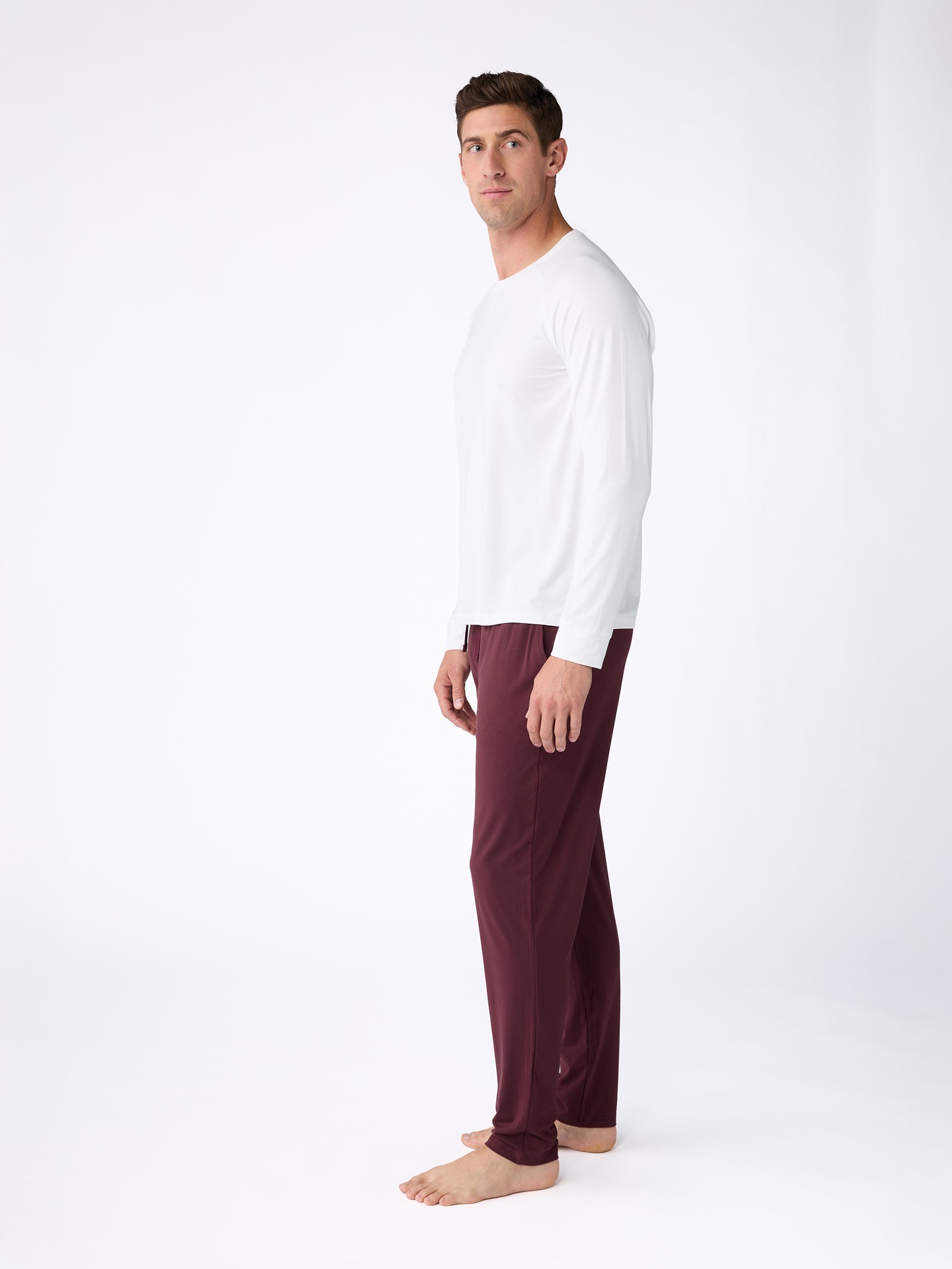 A man stands barefoot against a white background, wearing the Cozy Earth's Men's Stretch-Knit Bamboo Long Sleeve and maroon pants. His body is slightly angled to the side, and he is looking at the camera. 