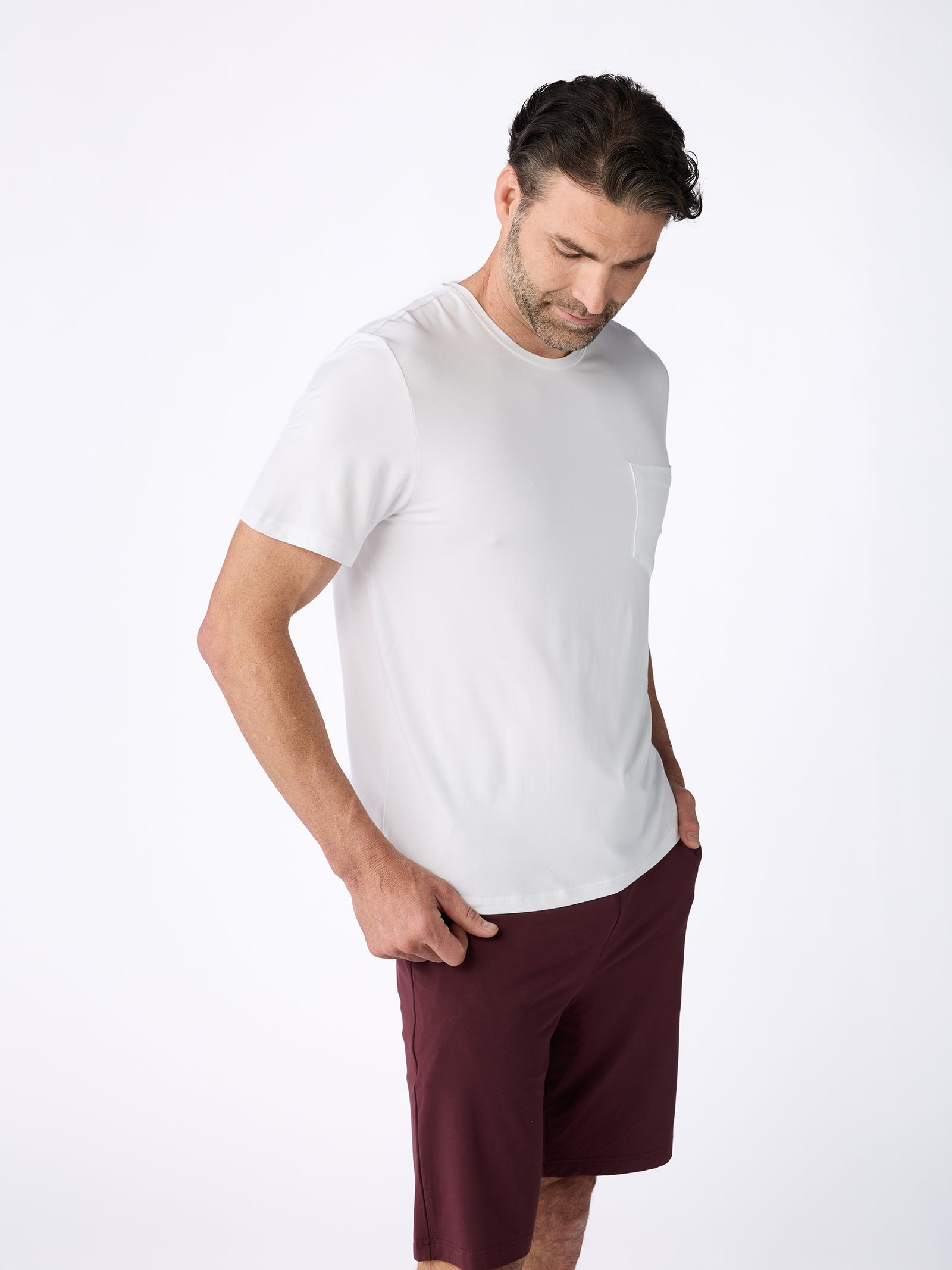 A man in a Men's Stretch-Knit Bamboo Lounge Tee by Cozy Earth and maroon shorts stands against a plain background, looking down with his hands in his pockets. 