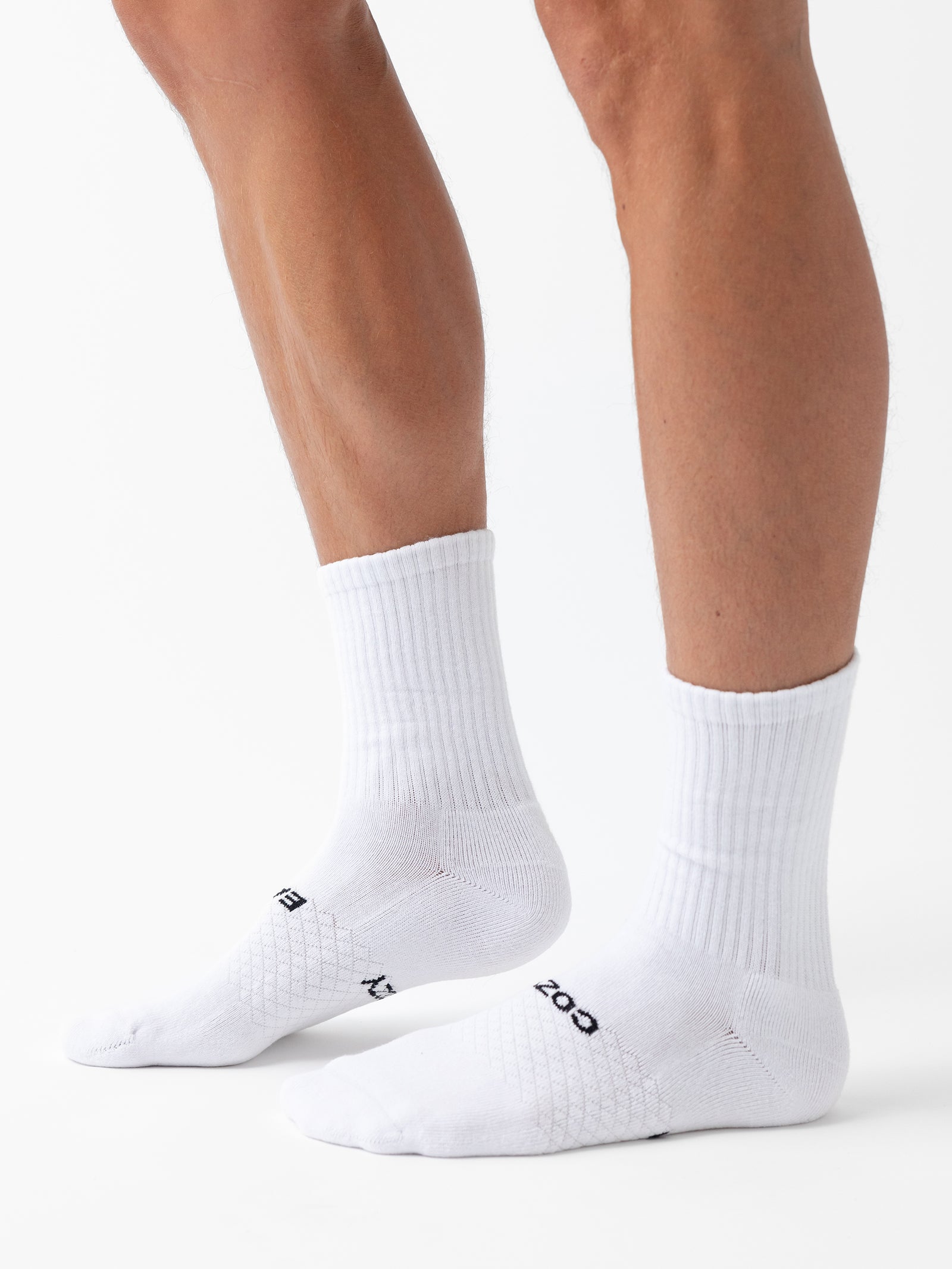 Close-up of a person's lower legs and feet wearing Cozy Earth's Essential Calf Socks. The white crew socks feature a ribbed texture around the ankle and have some text written on the sides near the toes. The background is white. 