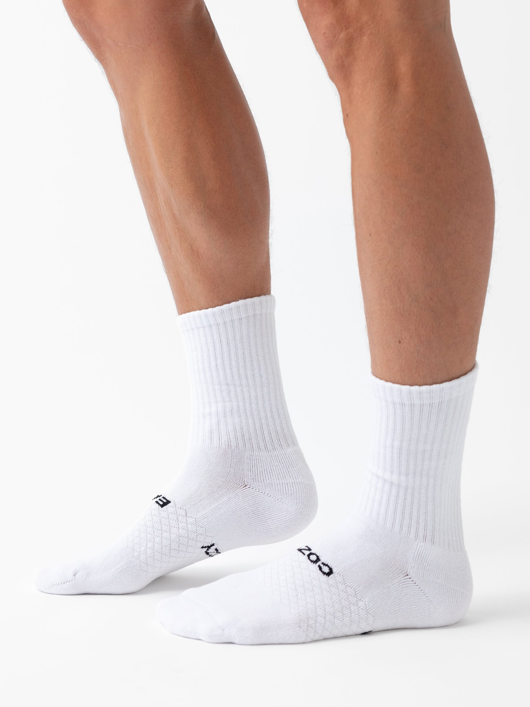 Two lower legs wearing Cozy Earth Essential Calf Socks stand on a white surface. The white socks feature ribbed cuffs, subtle textured patterns, and small black text near the toes. Sold as a 6-pack. 