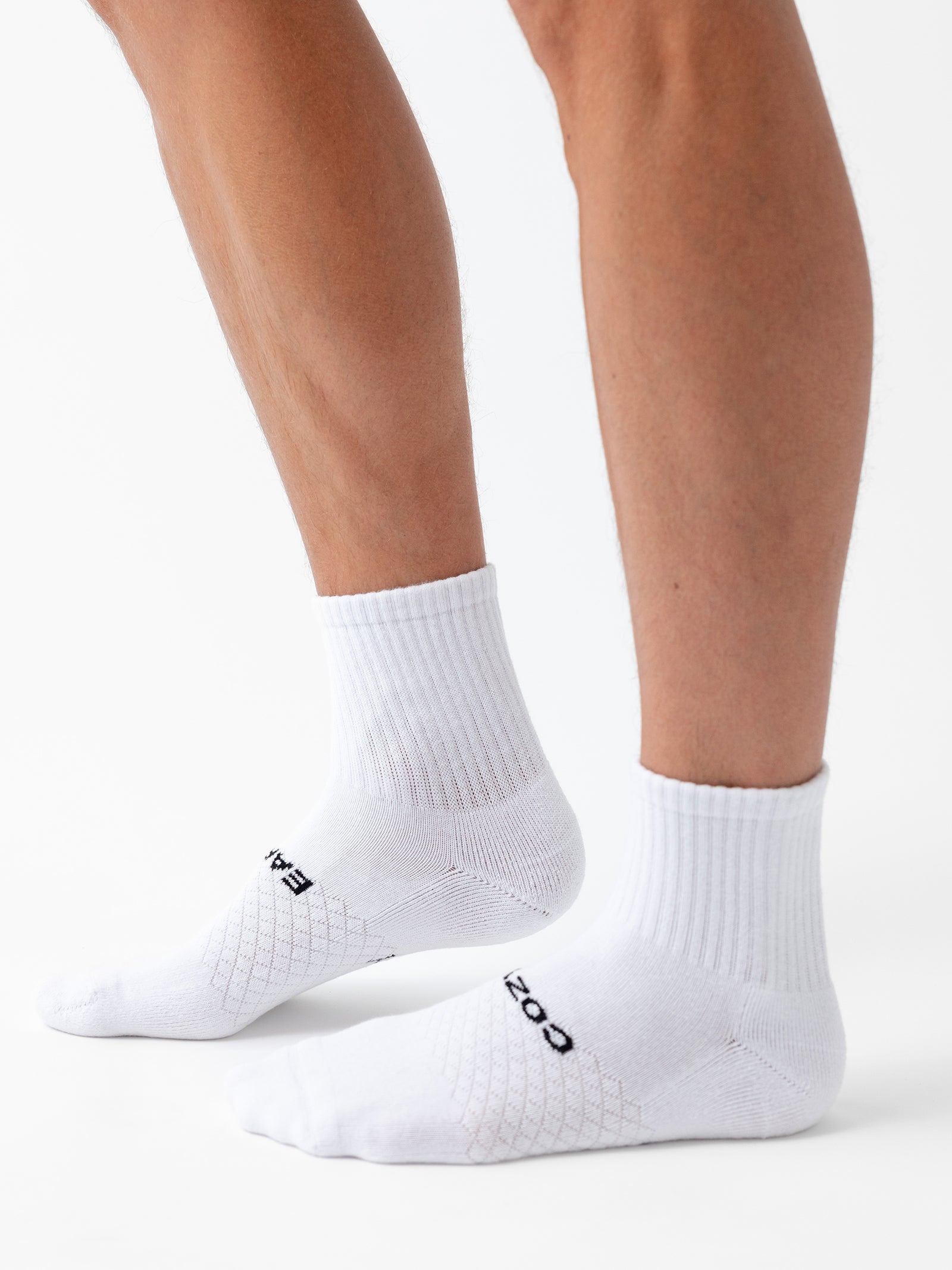 Two legs are shown wearing white Essential Quarter Socks from Cozy Earth’s 2-Pack, standing against a plain white background. The socks have a ribbed cuff with a grid-like texture and black text spelling “EAEO” on the top and “COM” on the bottom. 