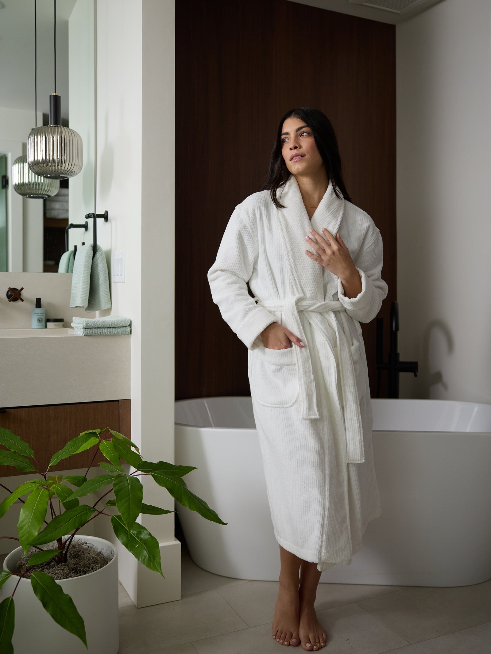 A person with long dark hair stands in a modern bathroom wearing a Cozy Earth Ribbed Terry Bath Robe. They are barefoot on a light-colored floor next to a large bathtub and a sink with a mirror and a plant. The room has a warm and minimalist vibe with wood and white accents. 