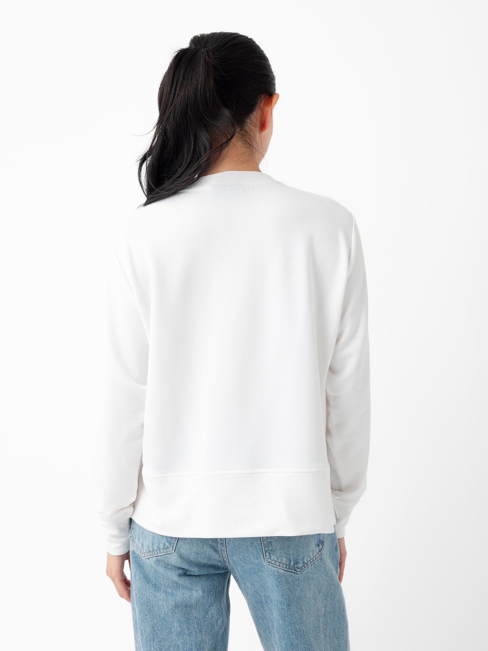 A person with long dark hair, standing with their back to the camera, is wearing a plain white Women's StudioLite Crewneck from Cozy Earth and blue jeans against a white background. 