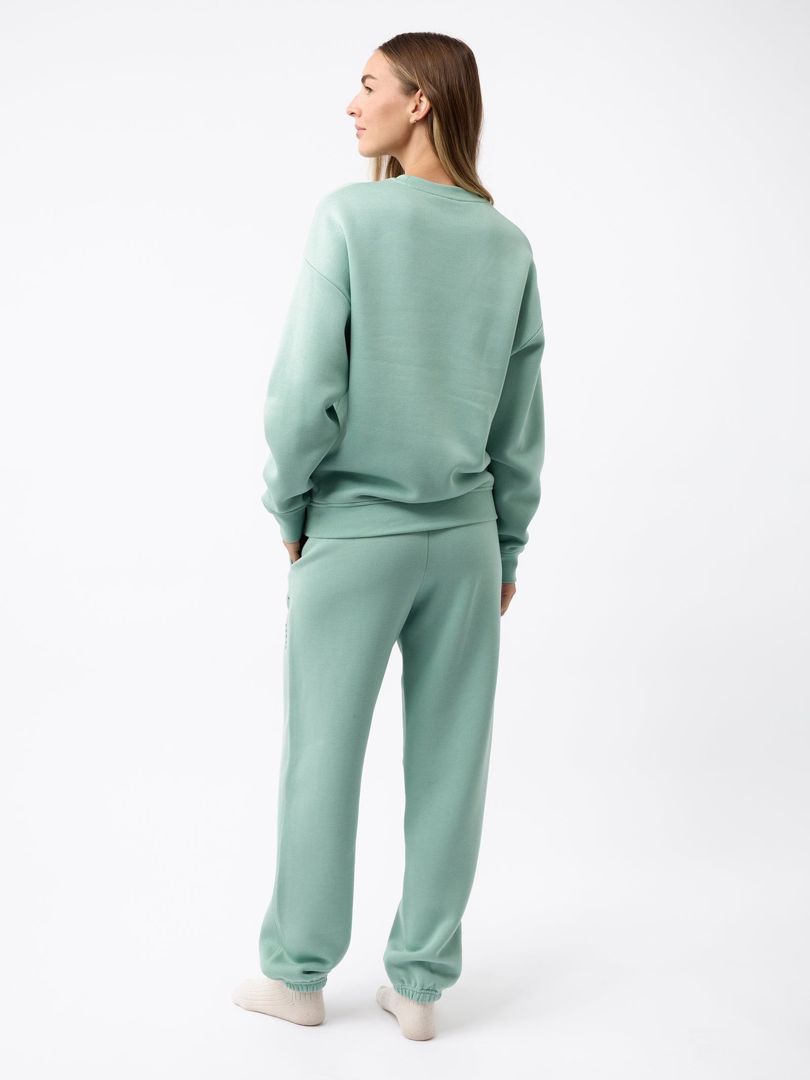 A person with long hair stands facing away, wearing the mint green Women's CityScape Crewneck and matching sweatpants from Cozy Earth. The outfit is casual and cozy, complemented by white sneakers, set against a plain white background. 