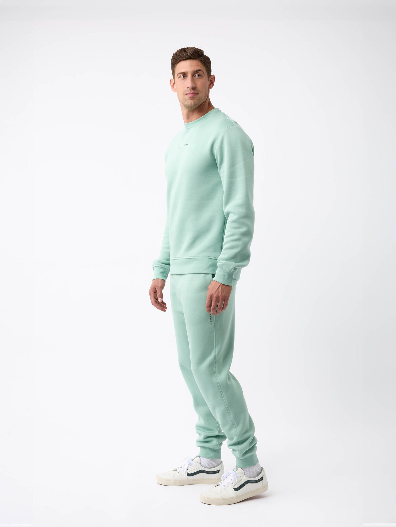 A man stands against a white background, wearing the Cozy Earth Men's CityScape Crewneck - Last Chance in light green with matching sweatpants and white sneakers, looking slightly left with a neutral expression.