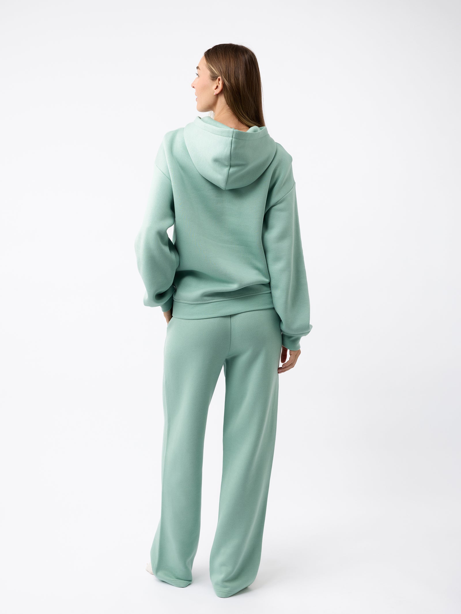 A person with long hair is wearing a green Women's CityScape Hoodie by Cozy Earth, along with matching pants. They are standing against a plain white background, facing away from the camera. 