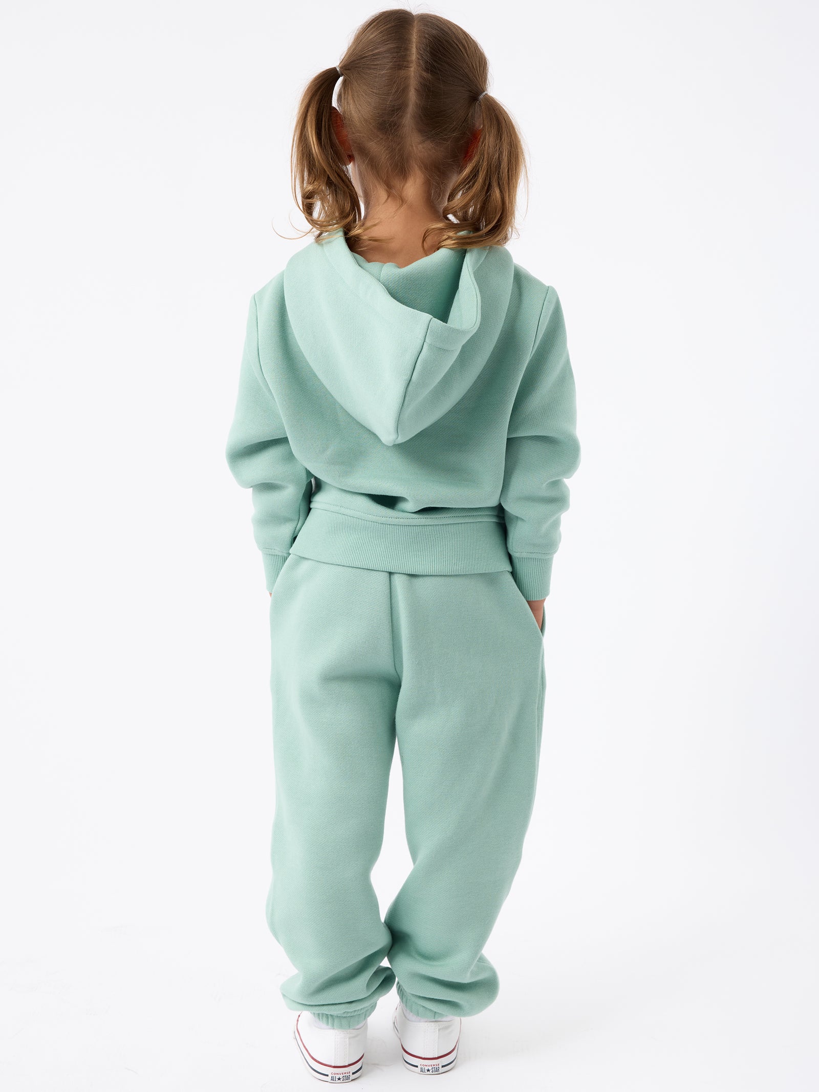 A young child with pigtails is facing away from the camera, dressed in a teal Kid's CityScape Hoodie by Cozy Earth, paired with matching pants. The child has their hands in their pockets and is wearing white sneakers against a plain white background. 