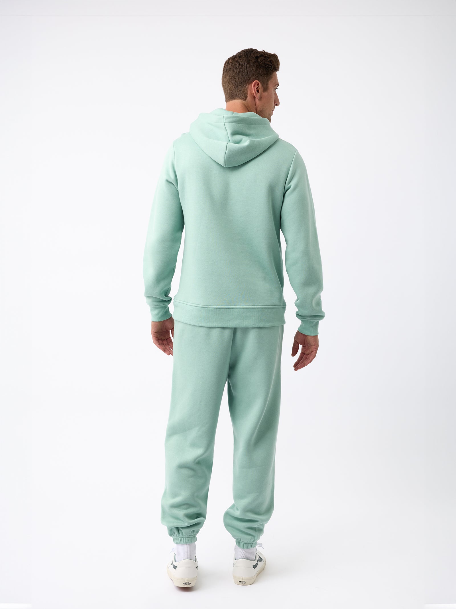 A person wearing a Men's CityScape Hoodie in light green from Cozy Earth, with matching sweatpants, stands facing away from the camera on a plain white background and is sporting white sneakers. 