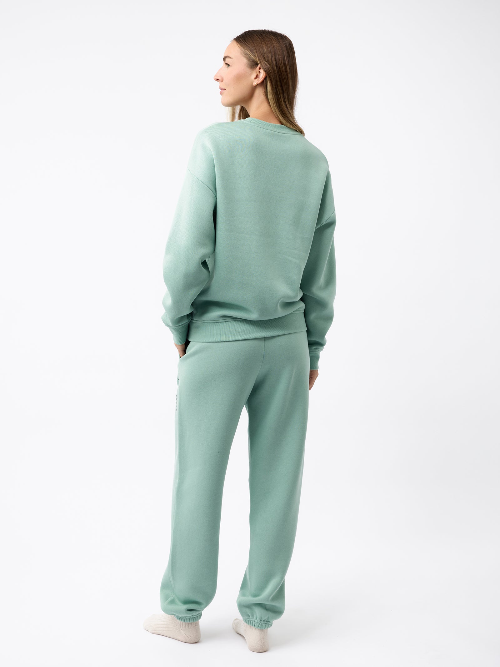 A person stands with their back to the camera, wearing a matching light teal sweatshirt and the Women's CityScape Sweatpant by Cozy Earth, complemented by white sneakers. The background is plain white. 